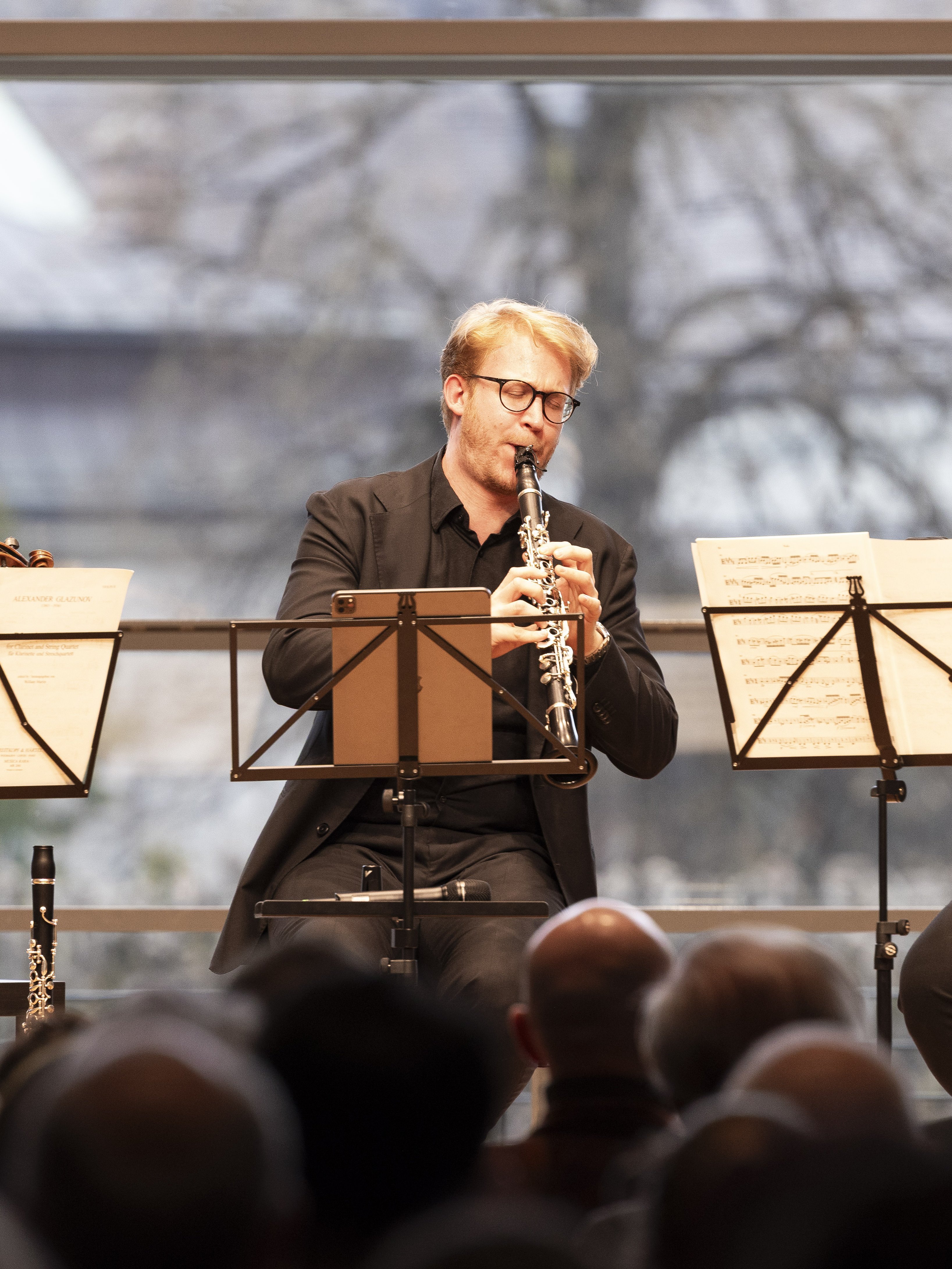 Ein Mann in einem schwarzen Anzug und mit Brille spielt auf der Bühne Klarinette und liest Noten. Er spielt vor einem Publikum, das ein großes Fenster mit Bäumen im Hintergrund sieht.