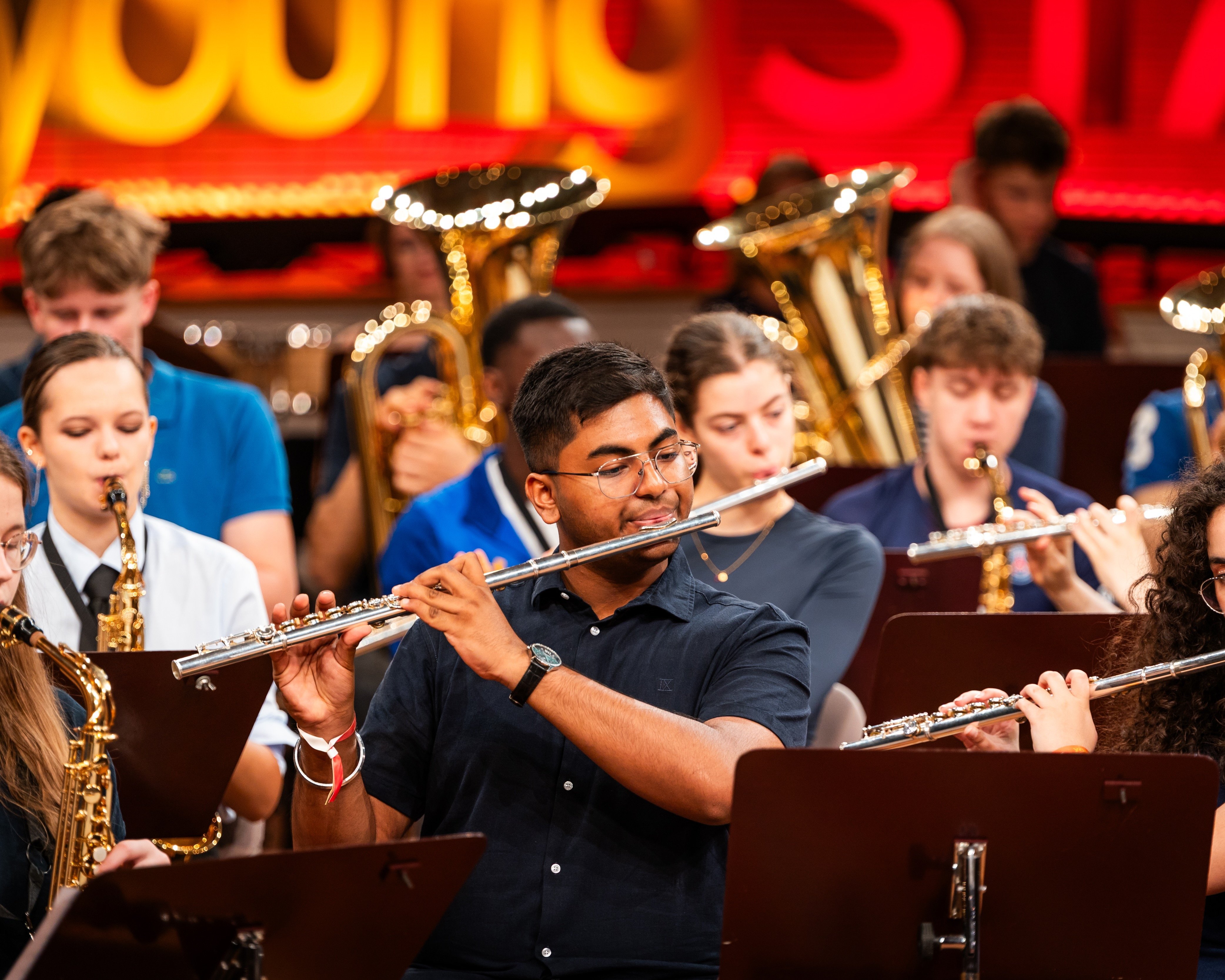A group of young musicians plays flutes and saxophones in an orchestra, with tubas in the background. They perform on stage in front of a brightly lit young STARS sign.