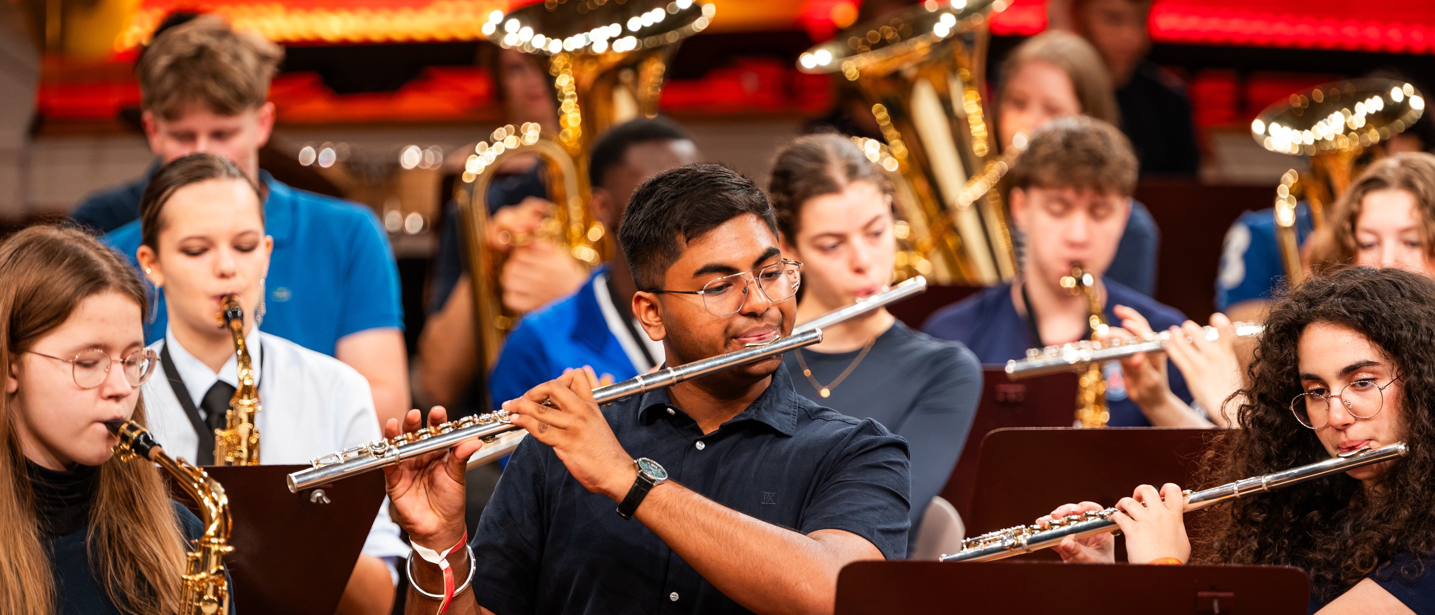 A group of young musicians plays flutes and saxophones in an orchestra, with tubas in the background. They perform on stage in front of a brightly lit young STARS sign.