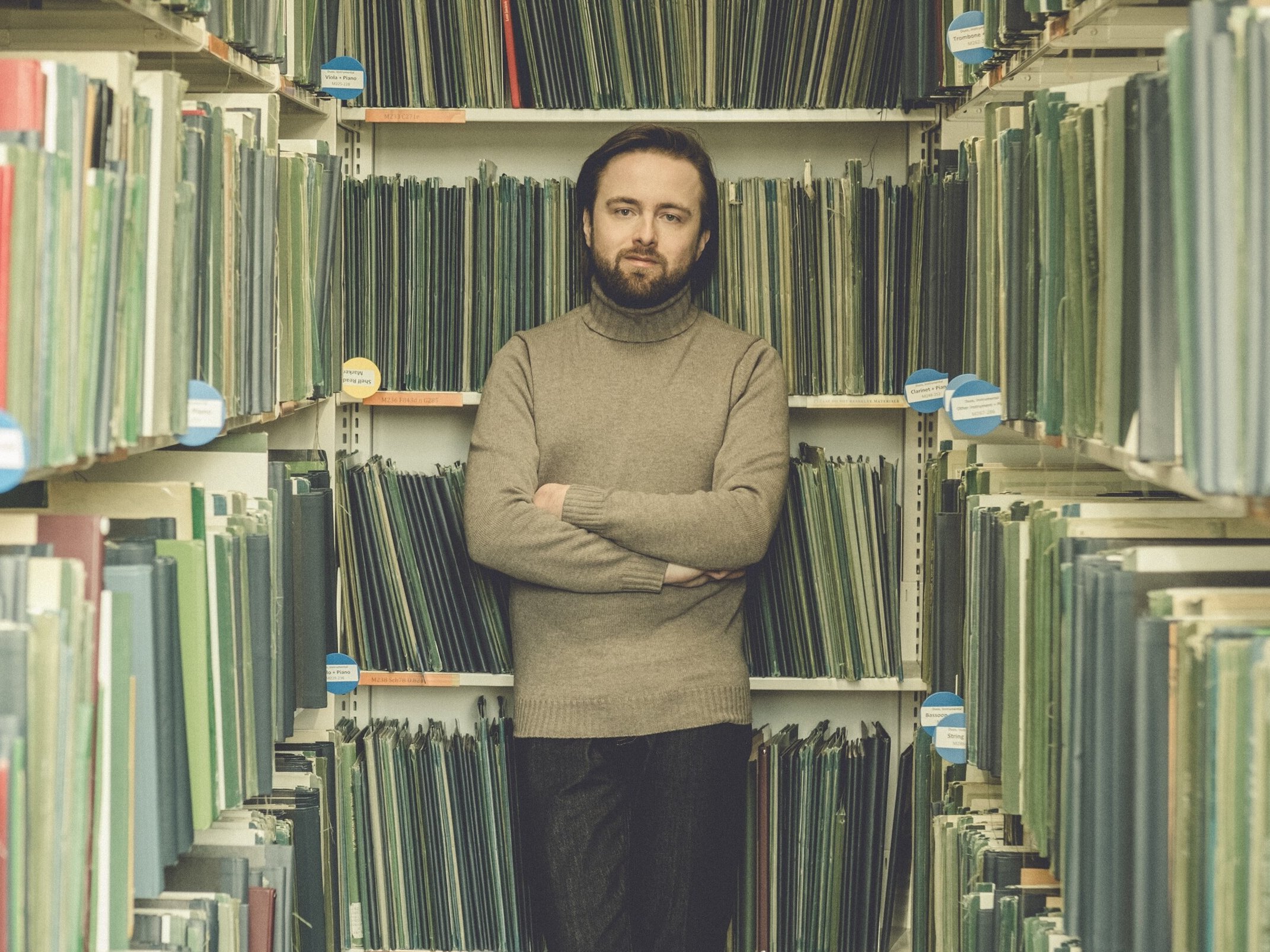 Ein bärtiger Daniil Trifonov steht mit verschränkten Armen zwischen Regalen mit grünen Ordnern in einer Bibliothek oder einem Archiv und blickt direkt in die Kamera.