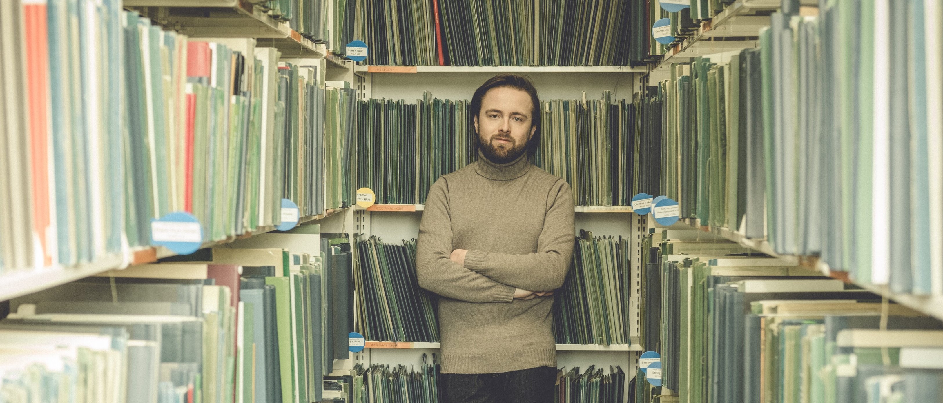 Ein bärtiger Daniil Trifonov steht mit verschränkten Armen zwischen Regalen mit grünen Ordnern in einer Bibliothek oder einem Archiv und blickt direkt in die Kamera.