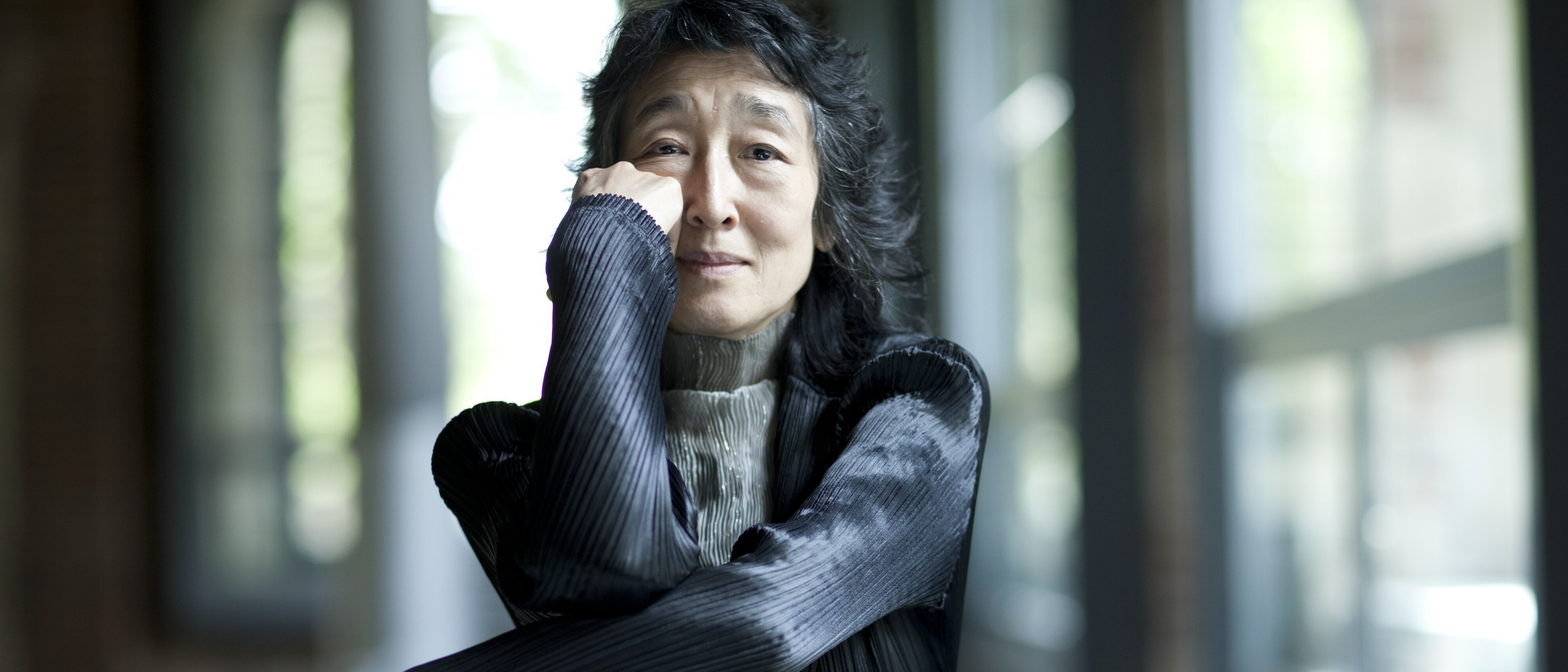 Mitsuko Uchida mit langen dunklen Haaren sitzt drinnen an großen Fenstern, stützt ihr Kinn auf die Hand und lächelt sanft. Sie trägt eine strukturierte schwarze Jacke und einen hellgrauen Rollkragenpullover.
