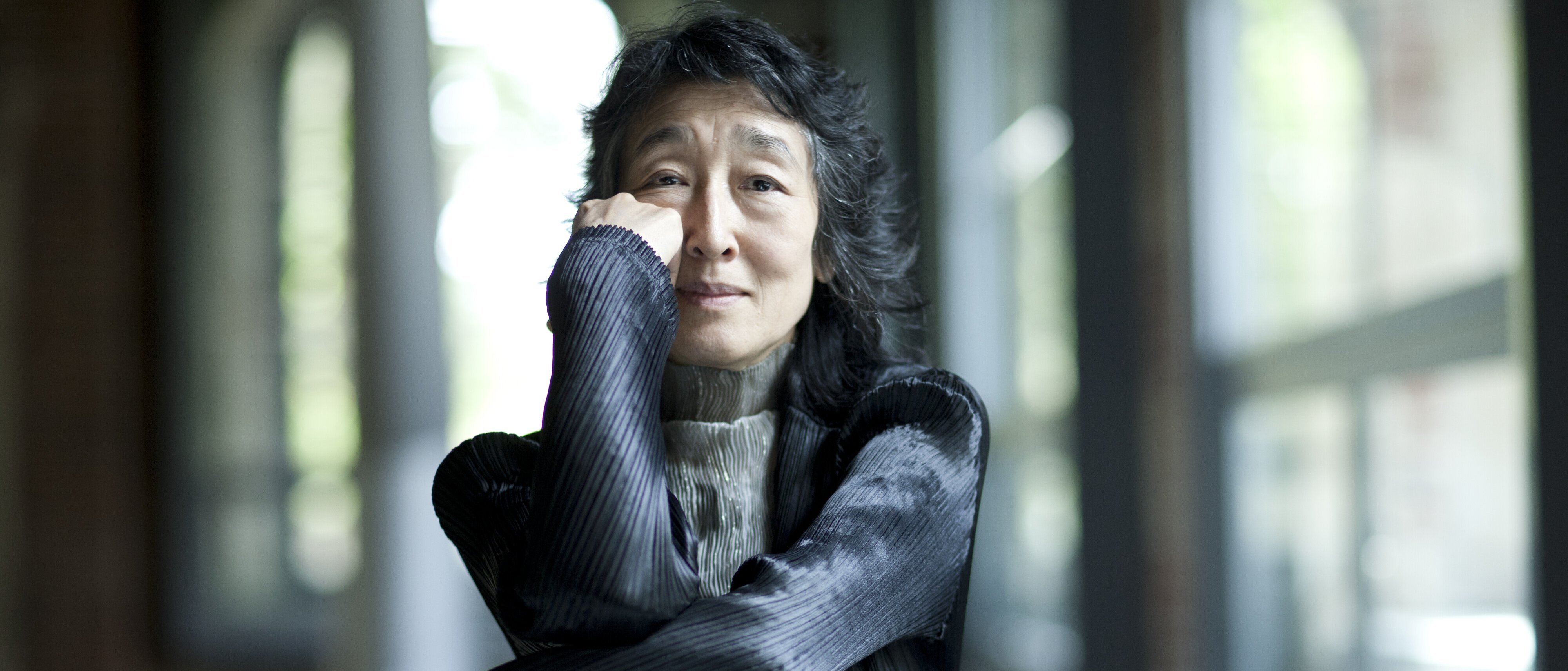 Mitsuko Uchida mit langen dunklen Haaren sitzt drinnen an großen Fenstern, stützt ihr Kinn auf die Hand und lächelt sanft. Sie trägt eine strukturierte schwarze Jacke und einen hellgrauen Rollkragenpullover.