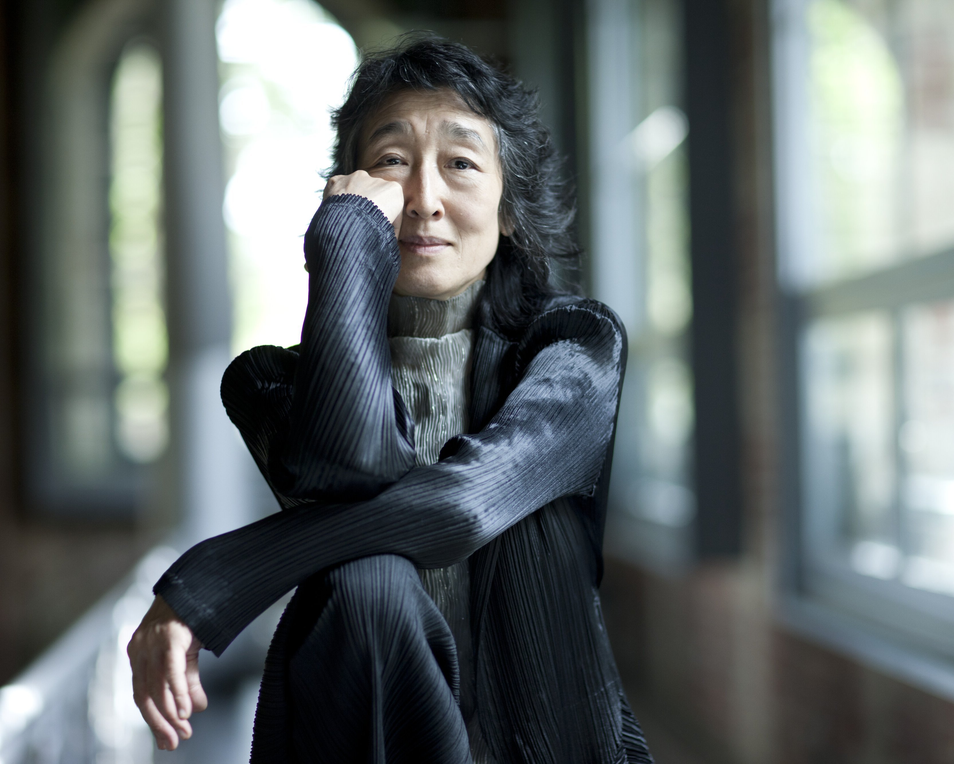 Mitsuko Uchida mit langen dunklen Haaren sitzt drinnen an großen Fenstern, stützt ihr Kinn auf die Hand und lächelt sanft. Sie trägt eine strukturierte schwarze Jacke und einen hellgrauen Rollkragenpullover.