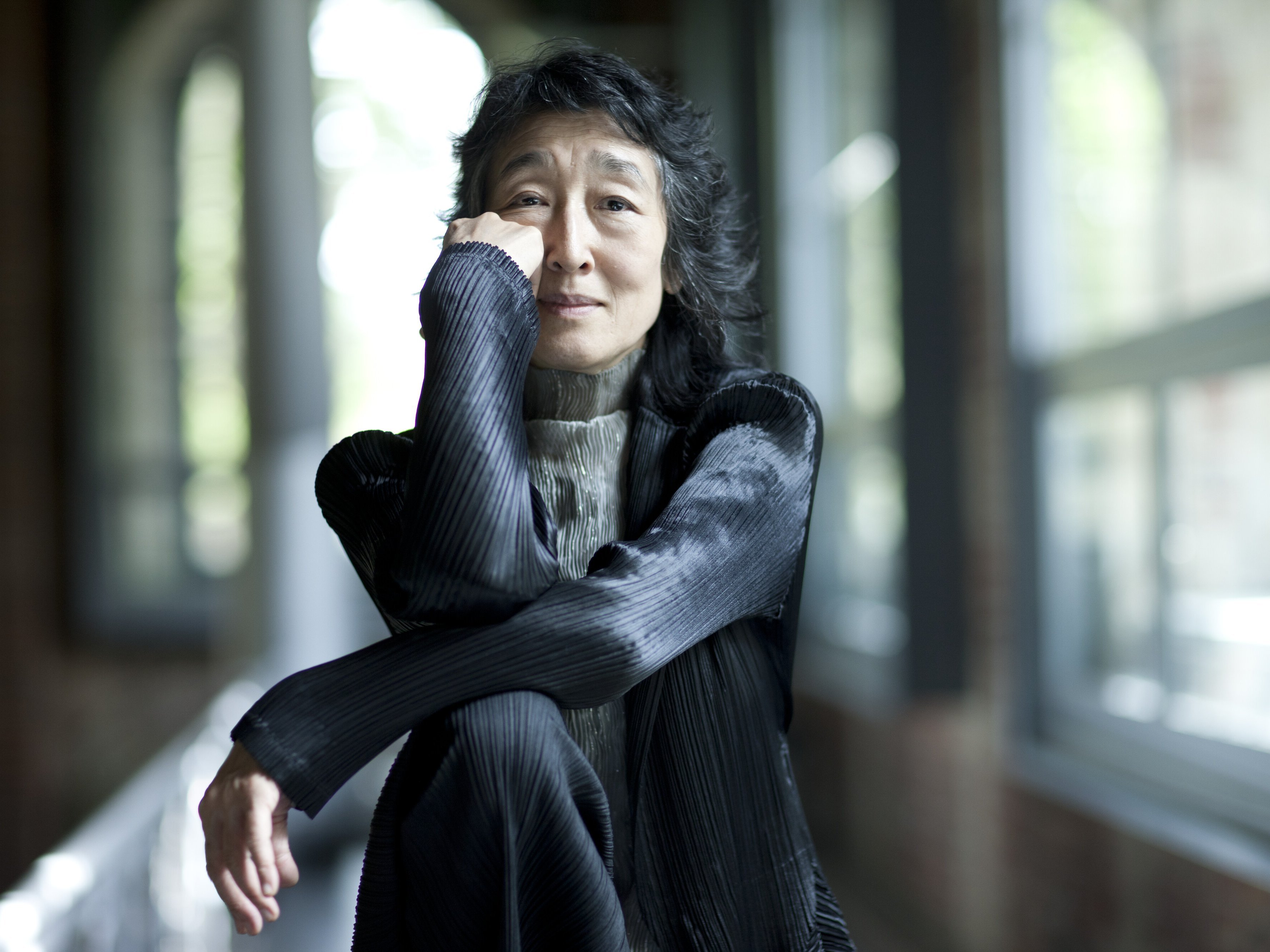 Mitsuko Uchida mit langen dunklen Haaren sitzt drinnen an großen Fenstern, stützt ihr Kinn auf die Hand und lächelt sanft. Sie trägt eine strukturierte schwarze Jacke und einen hellgrauen Rollkragenpullover.