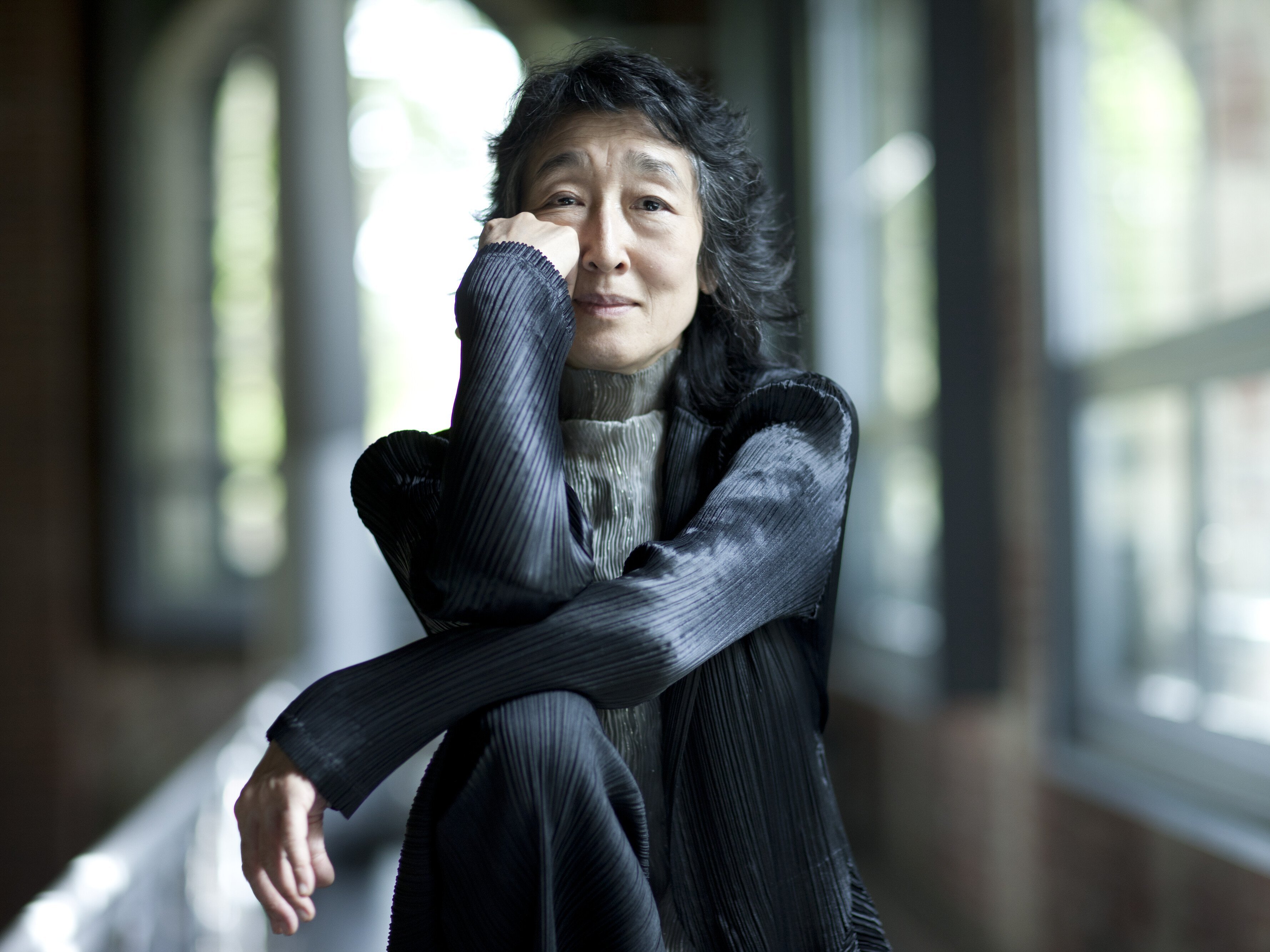 Mitsuko Uchida mit langen dunklen Haaren sitzt drinnen an großen Fenstern, stützt ihr Kinn auf die Hand und lächelt sanft. Sie trägt eine strukturierte schwarze Jacke und einen hellgrauen Rollkragenpullover.