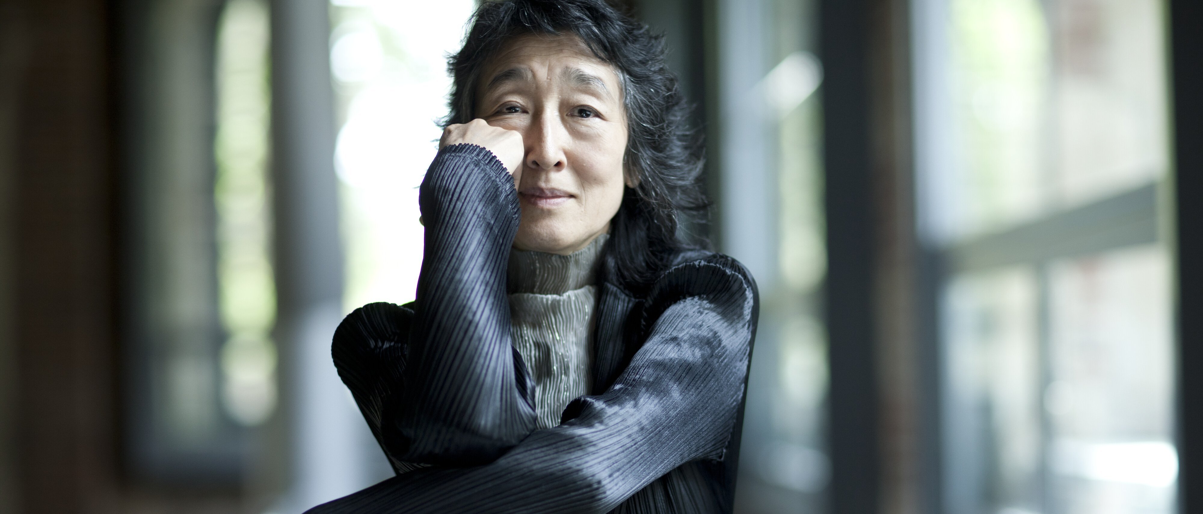 Mitsuko Uchida mit langen dunklen Haaren sitzt drinnen an großen Fenstern, stützt ihr Kinn auf die Hand und lächelt sanft. Sie trägt eine strukturierte schwarze Jacke und einen hellgrauen Rollkragenpullover.