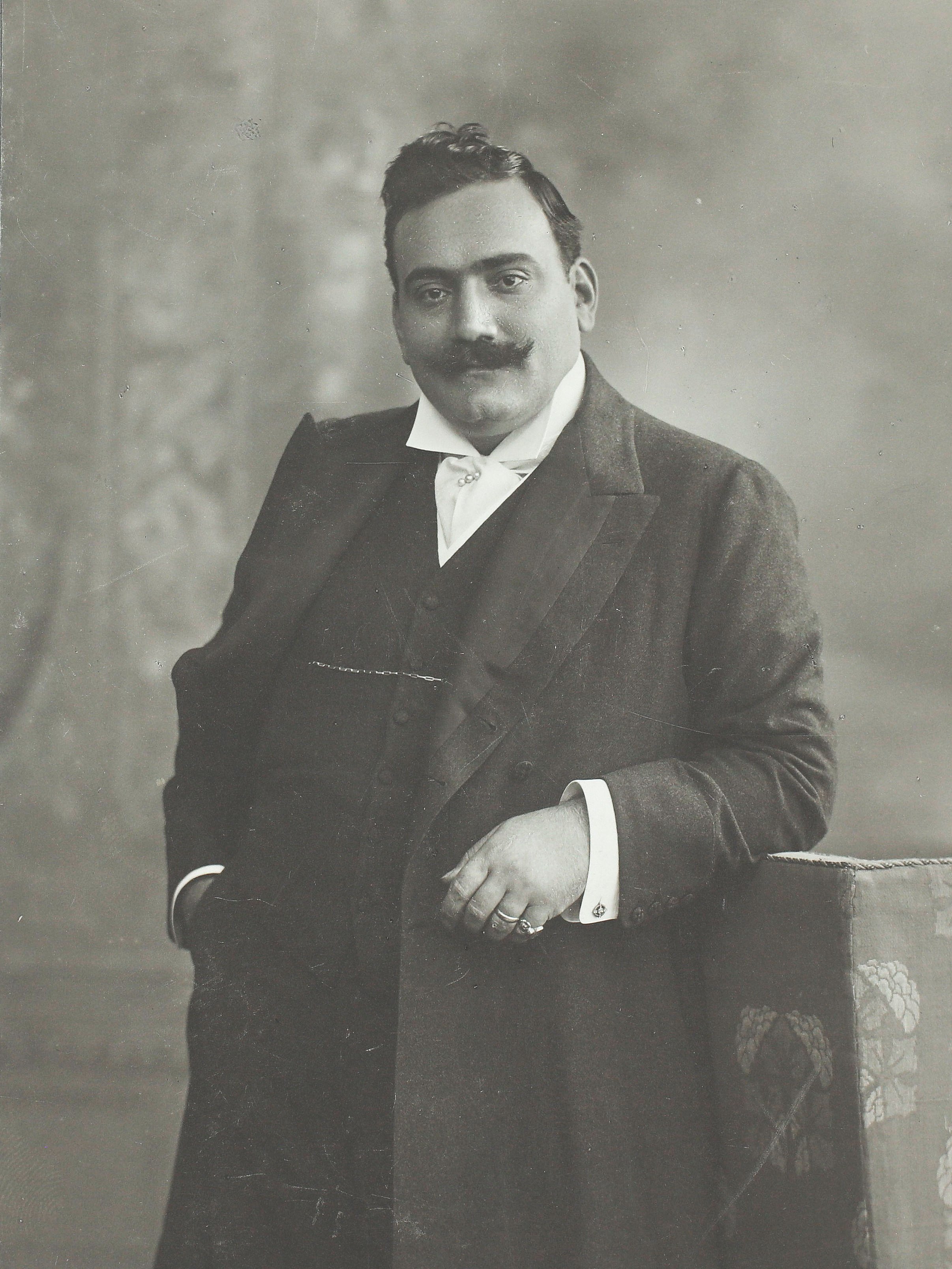 Ein Mann mit Schnurrbart in einem formellen Anzug posiert für ein klassisches Schwarz-Weiß-Porträt, wobei er eine Hand in der Tasche hat und die andere auf einer gemusterten Oberfläche ruht.