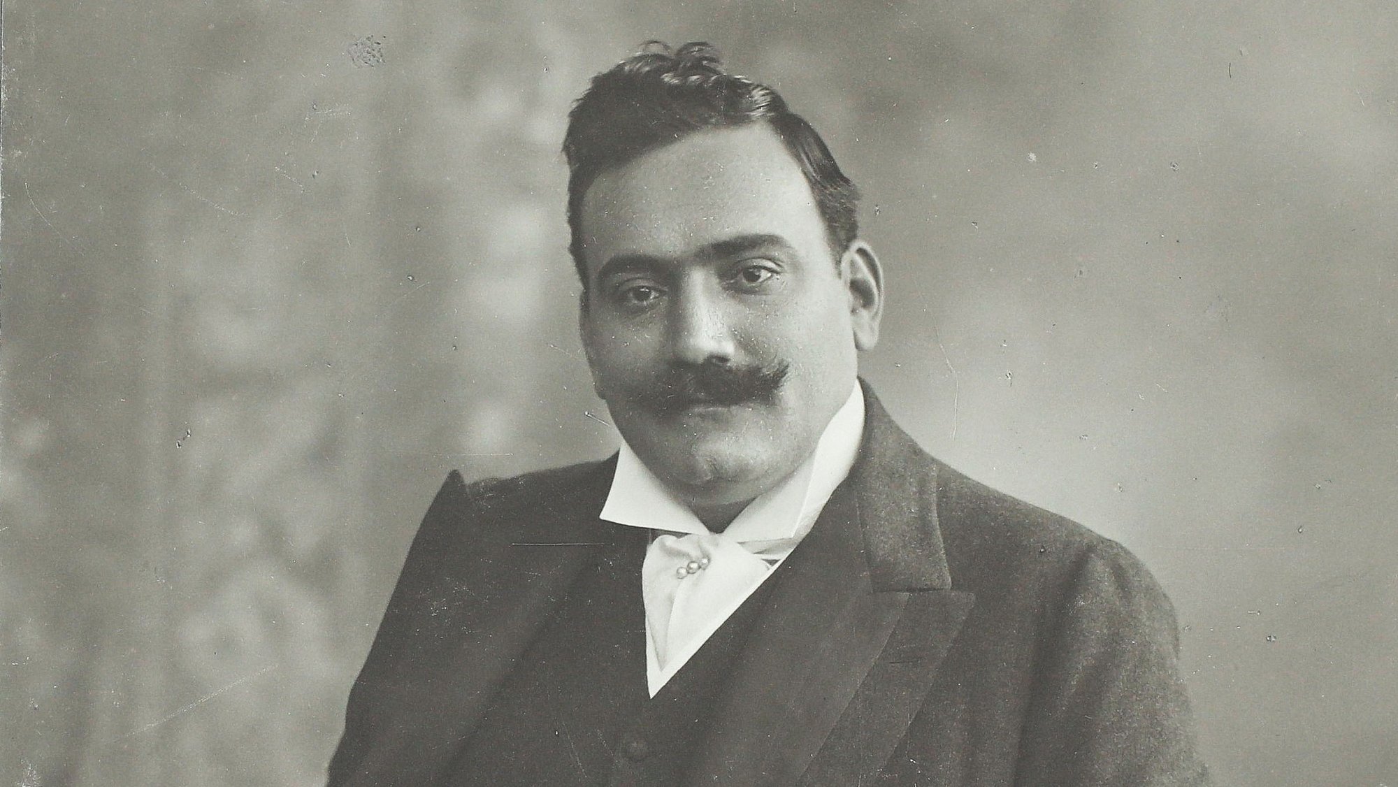 Ein Mann mit Schnurrbart in einem formellen Anzug posiert für ein klassisches Schwarz-Weiß-Porträt, wobei er eine Hand in der Tasche hat und die andere auf einer gemusterten Oberfläche ruht.