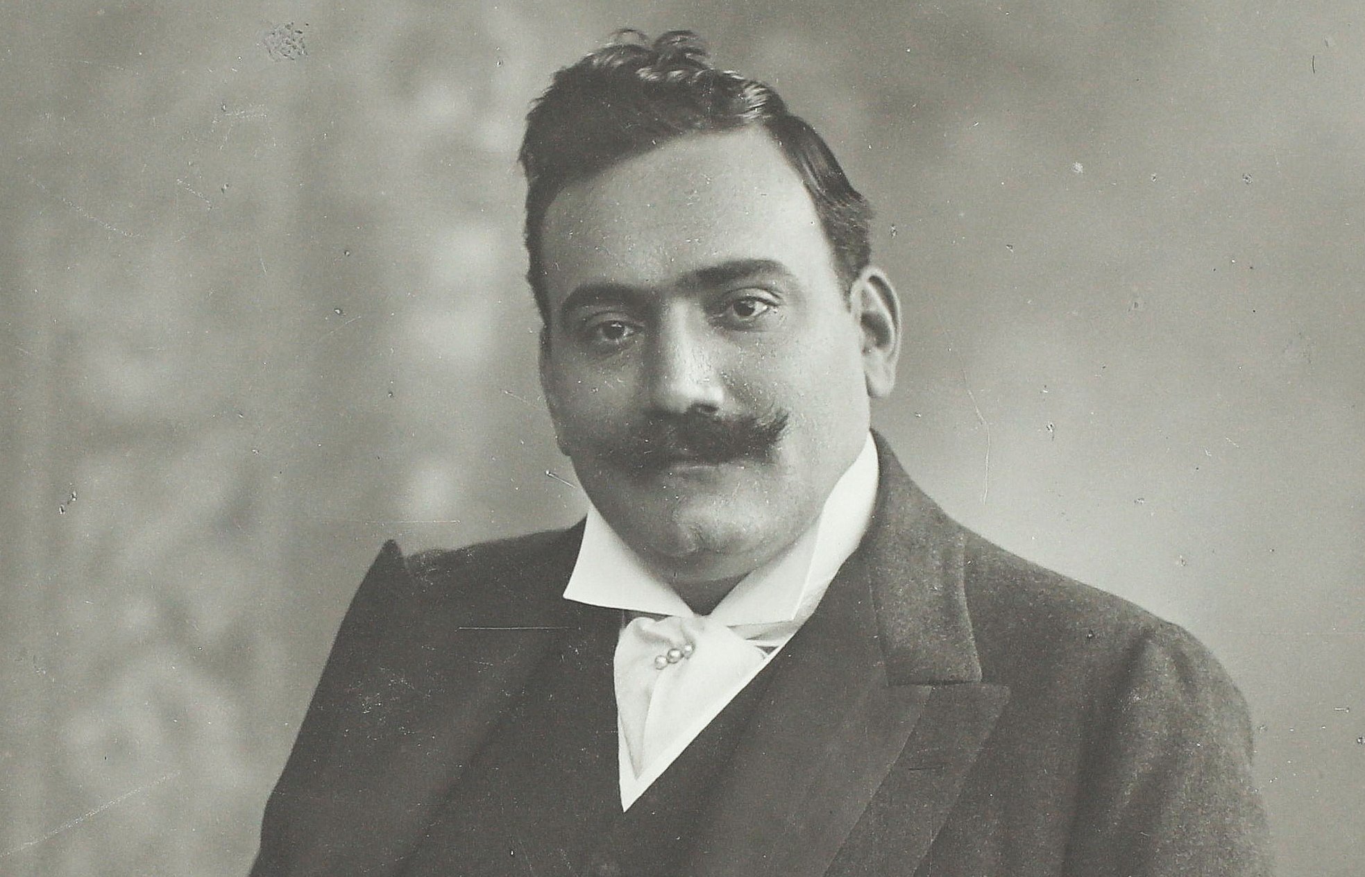 Ein Mann mit Schnurrbart in einem formellen Anzug posiert für ein klassisches Schwarz-Weiß-Porträt, wobei er eine Hand in der Tasche hat und die andere auf einer gemusterten Oberfläche ruht.