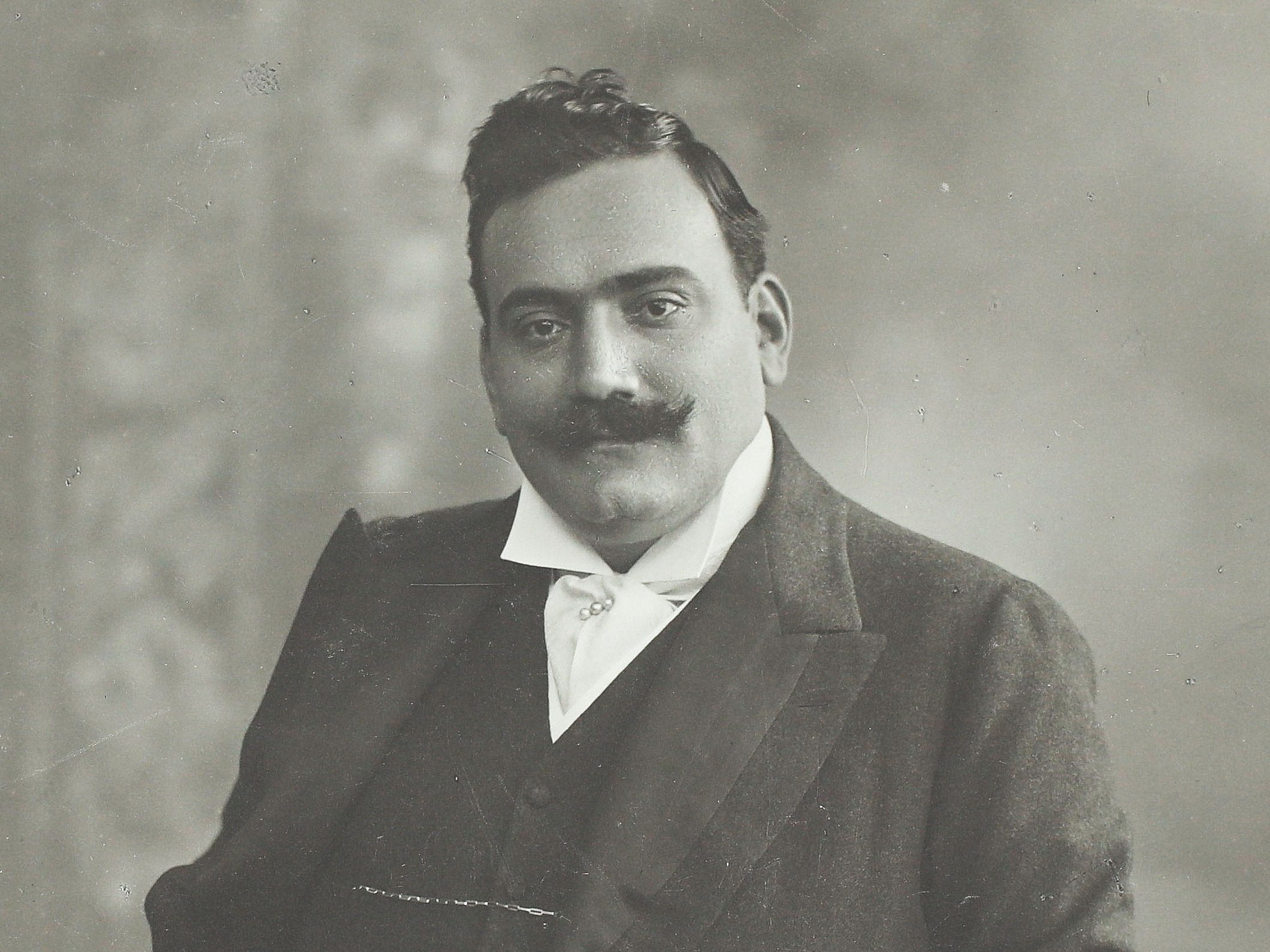 Ein Mann mit Schnurrbart in einem formellen Anzug posiert für ein klassisches Schwarz-Weiß-Porträt, wobei er eine Hand in der Tasche hat und die andere auf einer gemusterten Oberfläche ruht.