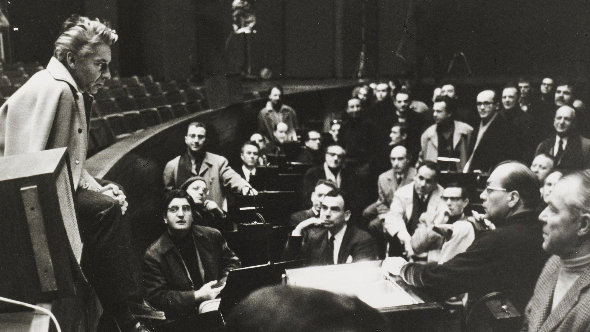 Karajan in einem hellen Mantel sitzt am Rande einer Bühne und spricht zu einer großen Gruppe von Menschen, die sich in einem Auditorium versammelt haben. Die meisten Menschen sind auf den Redner konzentriert, wodurch eine engagierte Atmosphäre entsteht.