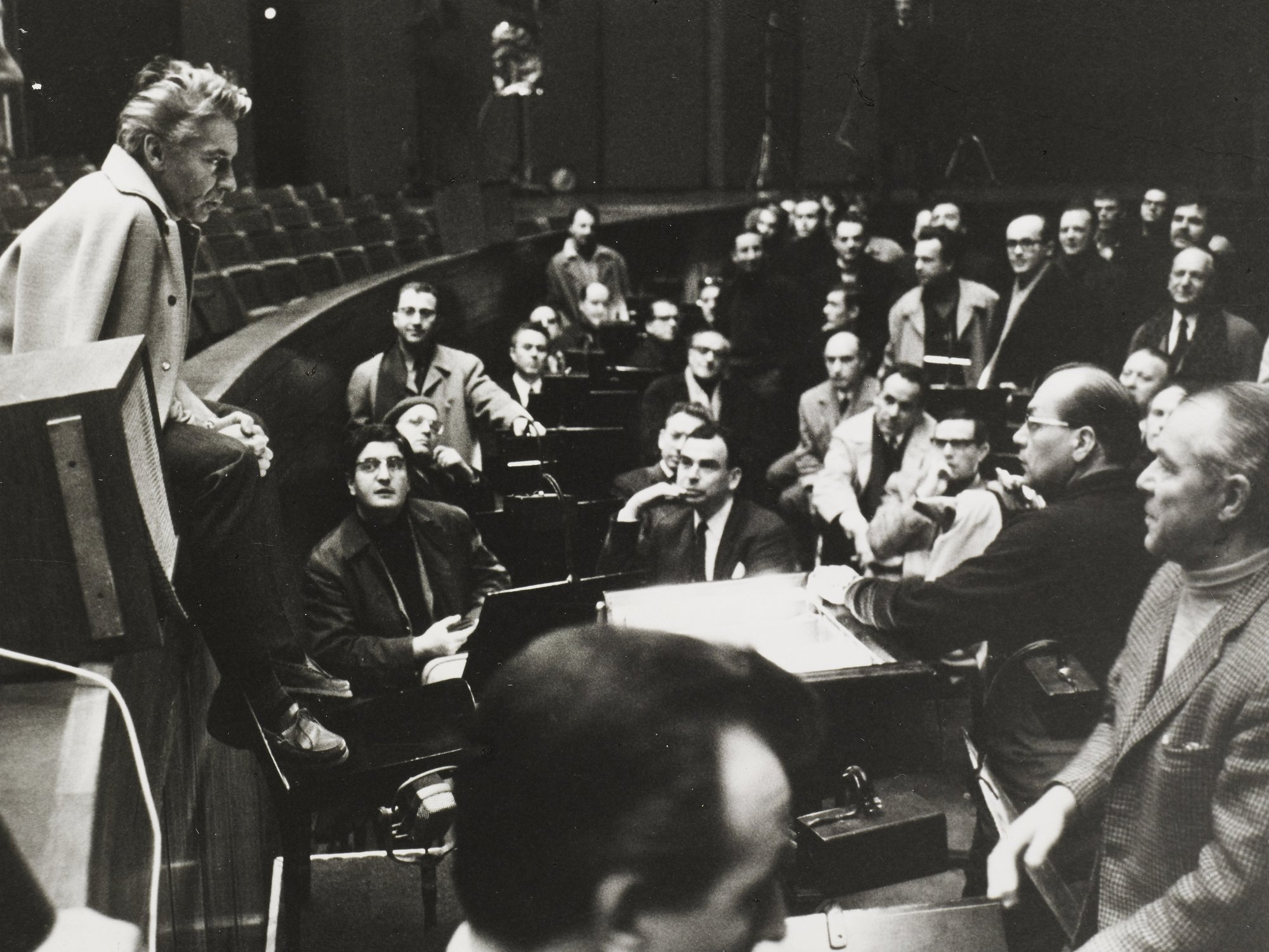 A group of people gathers in an auditorium, attentively listening to karajan seated casually on the edge of a platform at the front. The mood appears focused and engaged.