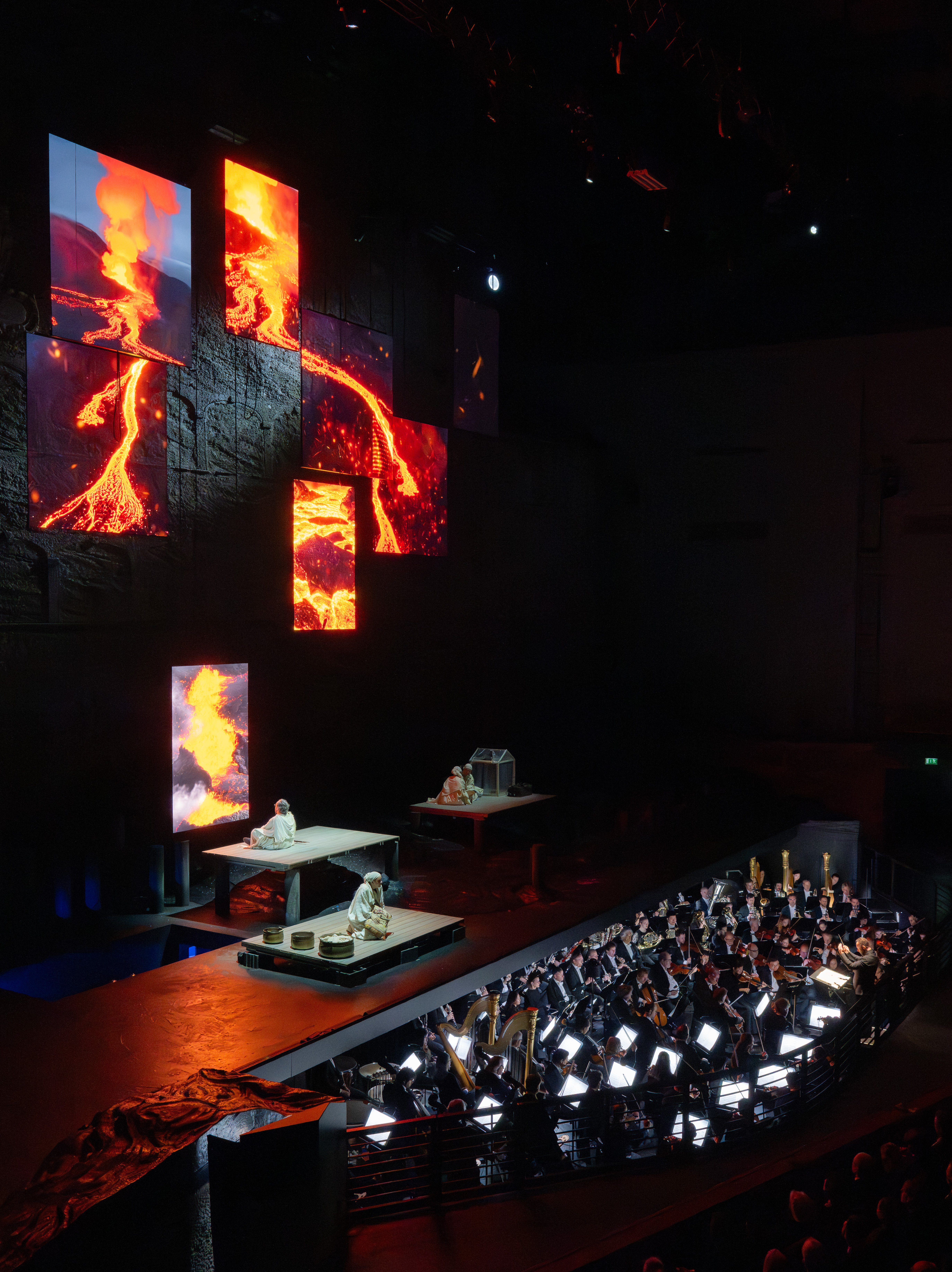 Eine Bühne mit Darstellern befindet sich über einem Orchestergraben, während große vertikale Bildschirme lebendige Bilder von fließender Lava zeigen, die eine dramatische Kulisse in dem schwach beleuchteten Theater bilden.