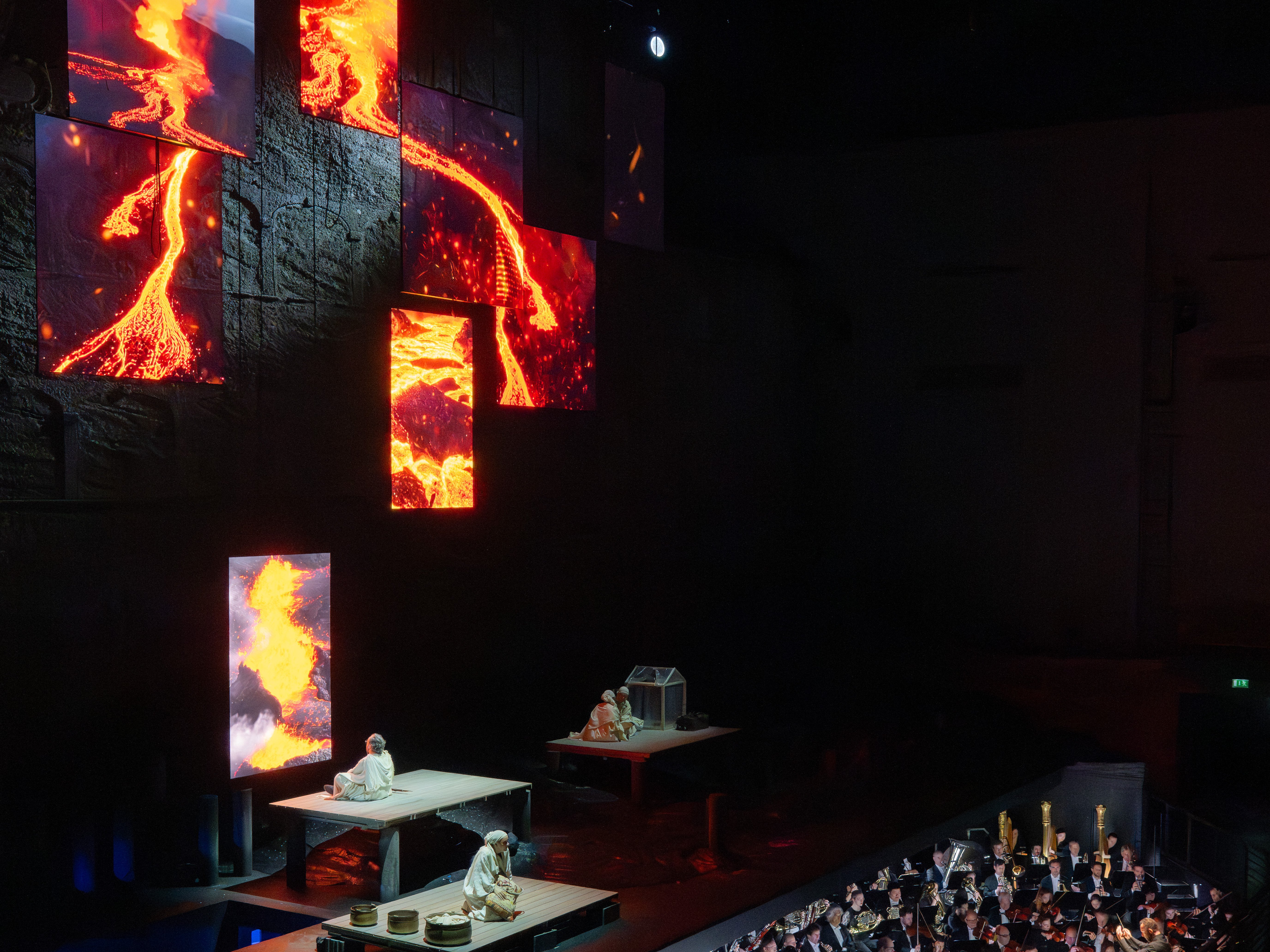 Eine Bühne mit Darstellern befindet sich über einem Orchestergraben, während große vertikale Bildschirme lebendige Bilder von fließender Lava zeigen, die eine dramatische Kulisse in dem schwach beleuchteten Theater bilden.