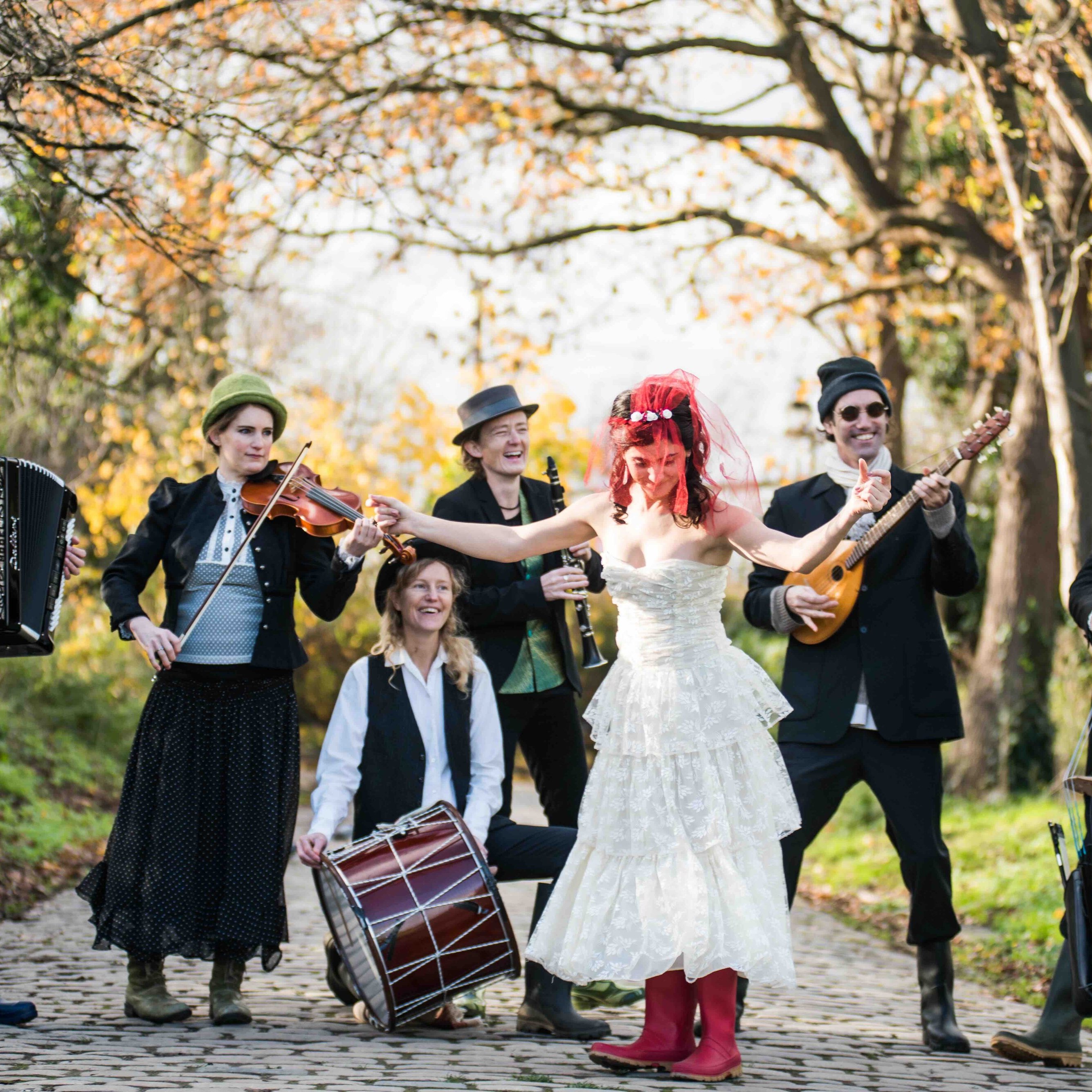 Eine Gruppe von sechs Musikern spielt im Freien auf einem Kopfsteinpflasterweg, im Hintergrund sind herbstlich gefärbte Bäume zu sehen. Eine Person trägt ein weißes Kleid und rote Stiefel, hält eine Gitarre in der Hand und singt.