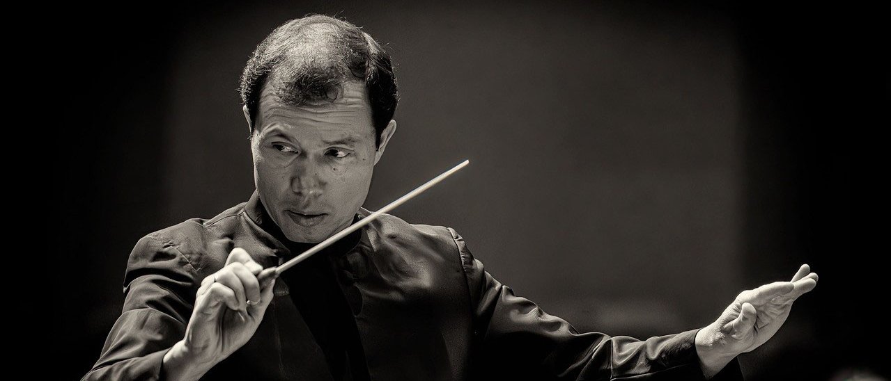 Ein Dirigent in formeller Kleidung leitet ein Orchester, hält in der einen Hand einen Taktstock und gestikuliert mit der anderen, ganz auf die Musik konzentriert. Der Hintergrund ist dunkel und unscharf.