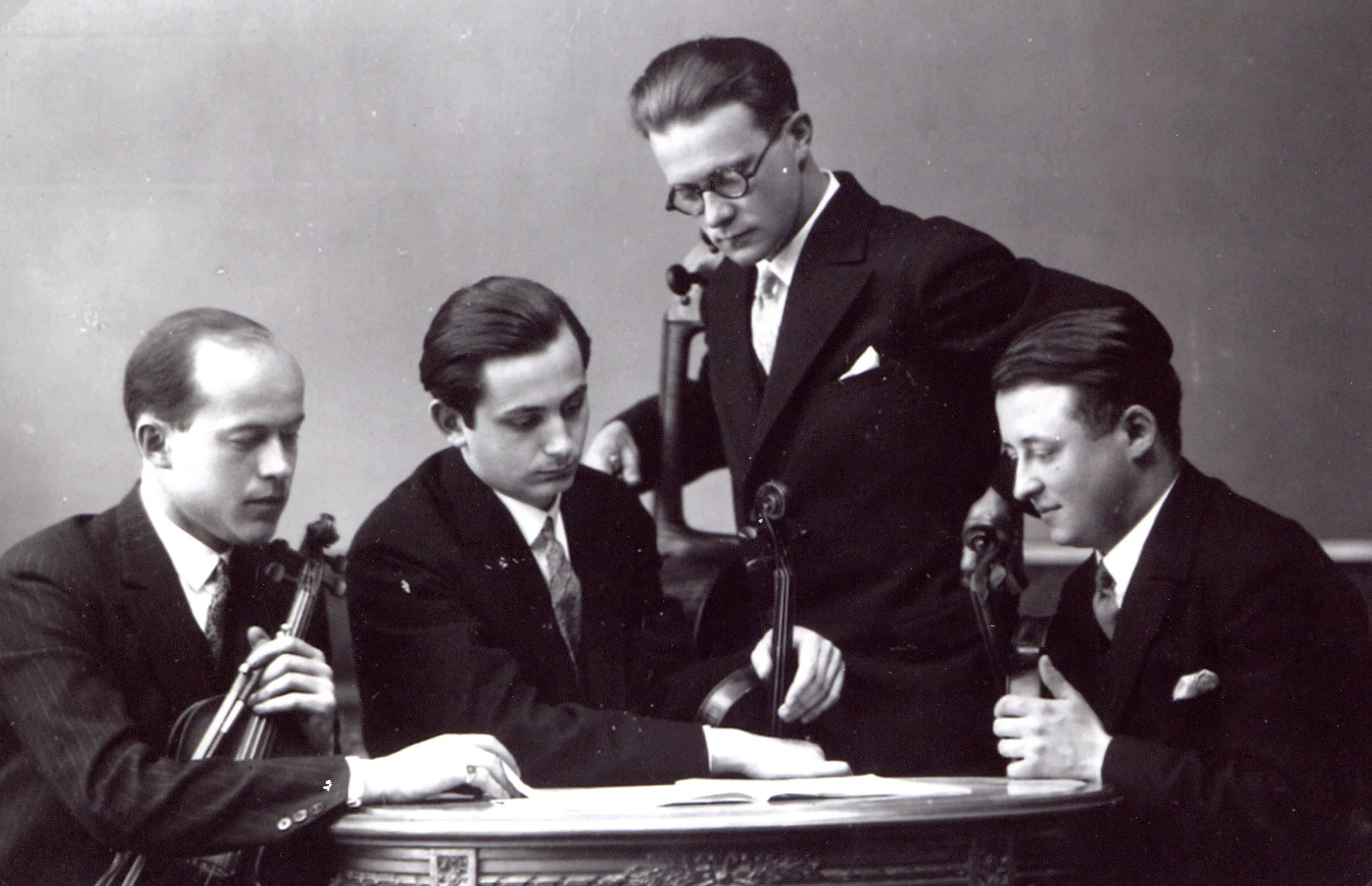 Vier Männer in Anzügen sitzen und stehen um einen kleinen runden Tisch herum und sehen sich Notenblätter an. Drei halten Geigen und einer ein Cello, was darauf hindeutet, dass es sich um klassische Musiker handelt, die sich auf einen Auftritt vorbereiten.