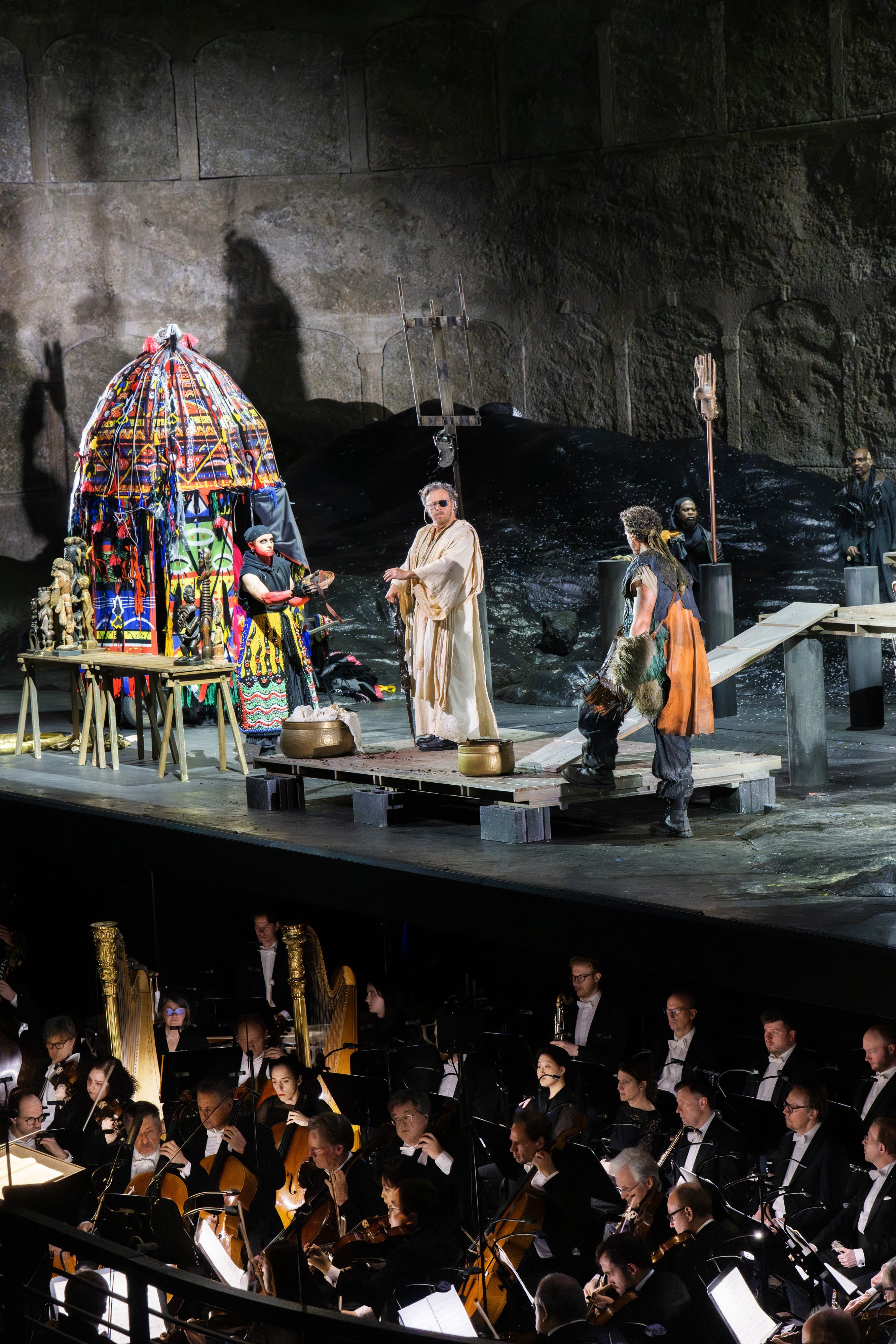 A theatrical performance onstage with actors in colorful costumes near a vibrant structure, while an orchestra plays below in the orchestra pit. The set has a dramatic, rocky background.