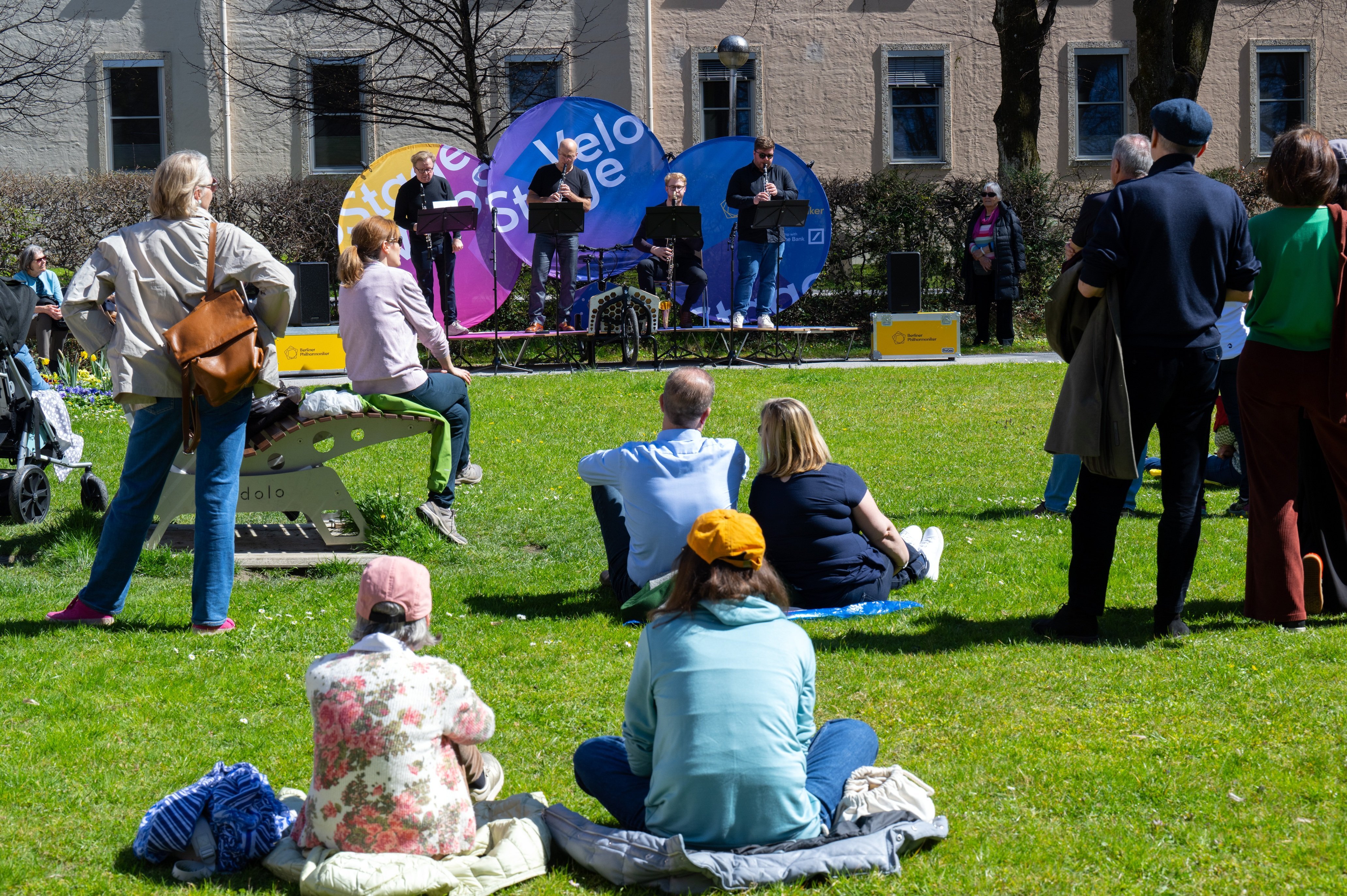 People sit and stand on grass in a park, watching a small outdoor stage with four speakers in front of colorful banners. The weather is sunny and bright.
