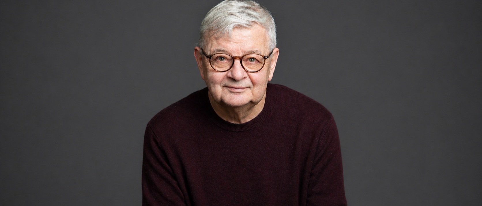 Ein älterer Mann mit kurzen grauen Haaren und einer runden Brille, der einen dunkelkastanienbraunen Pullover trägt, sitzt und lächelt sanft vor einem schlichten dunkelgrauen Hintergrund.