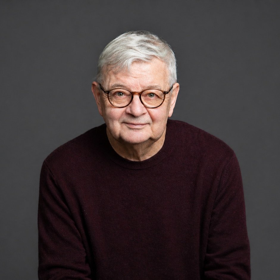 Ein älterer Mann mit kurzen grauen Haaren und einer runden Brille, der einen dunkelkastanienbraunen Pullover trägt, sitzt und lächelt sanft vor einem schlichten dunkelgrauen Hintergrund.