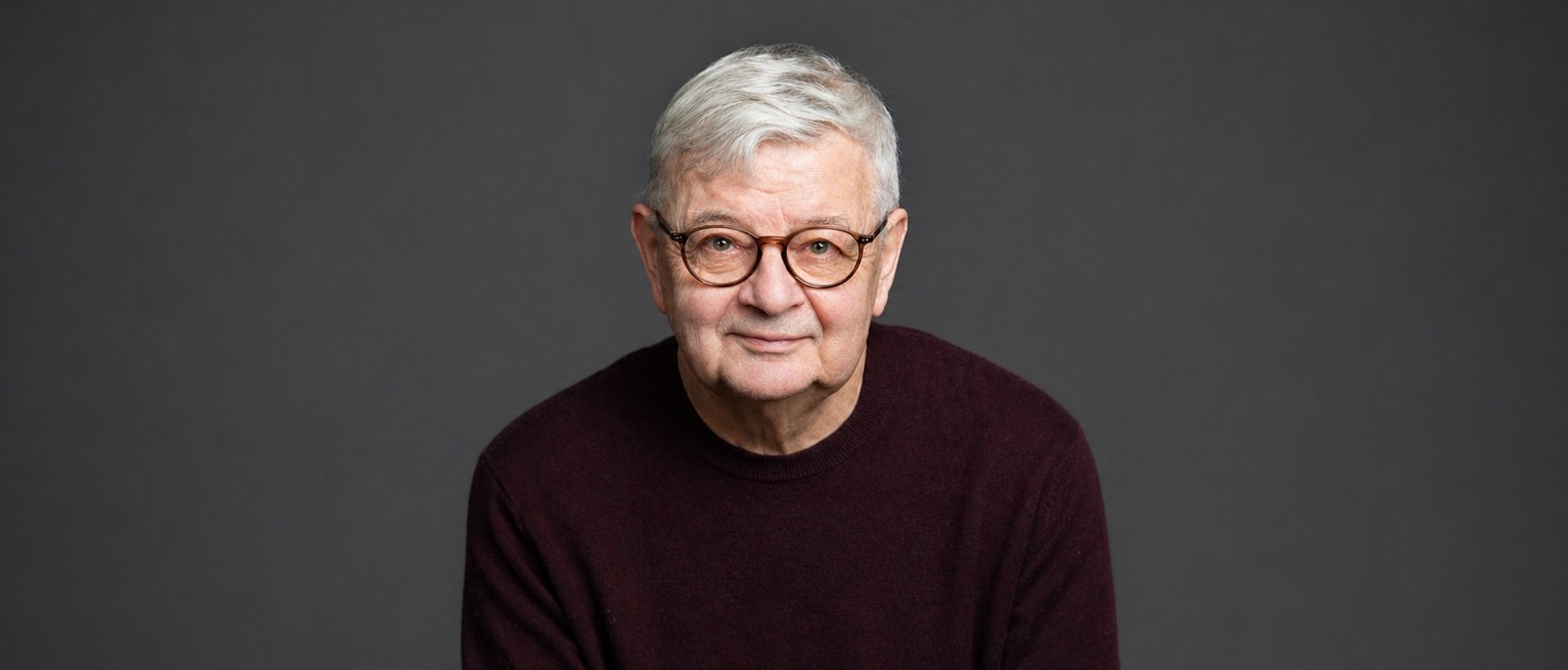 Ein älterer Mann mit kurzen grauen Haaren und einer runden Brille, der einen dunkelkastanienbraunen Pullover trägt, sitzt und lächelt sanft vor einem schlichten dunkelgrauen Hintergrund.