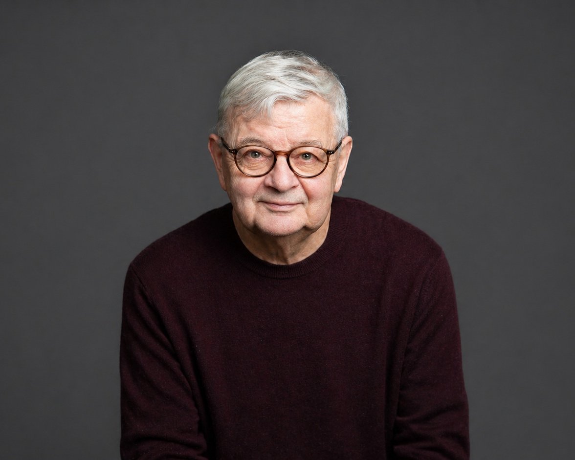 Ein älterer Mann mit kurzen grauen Haaren und einer runden Brille, der einen dunkelkastanienbraunen Pullover trägt, sitzt und lächelt sanft vor einem schlichten dunkelgrauen Hintergrund.