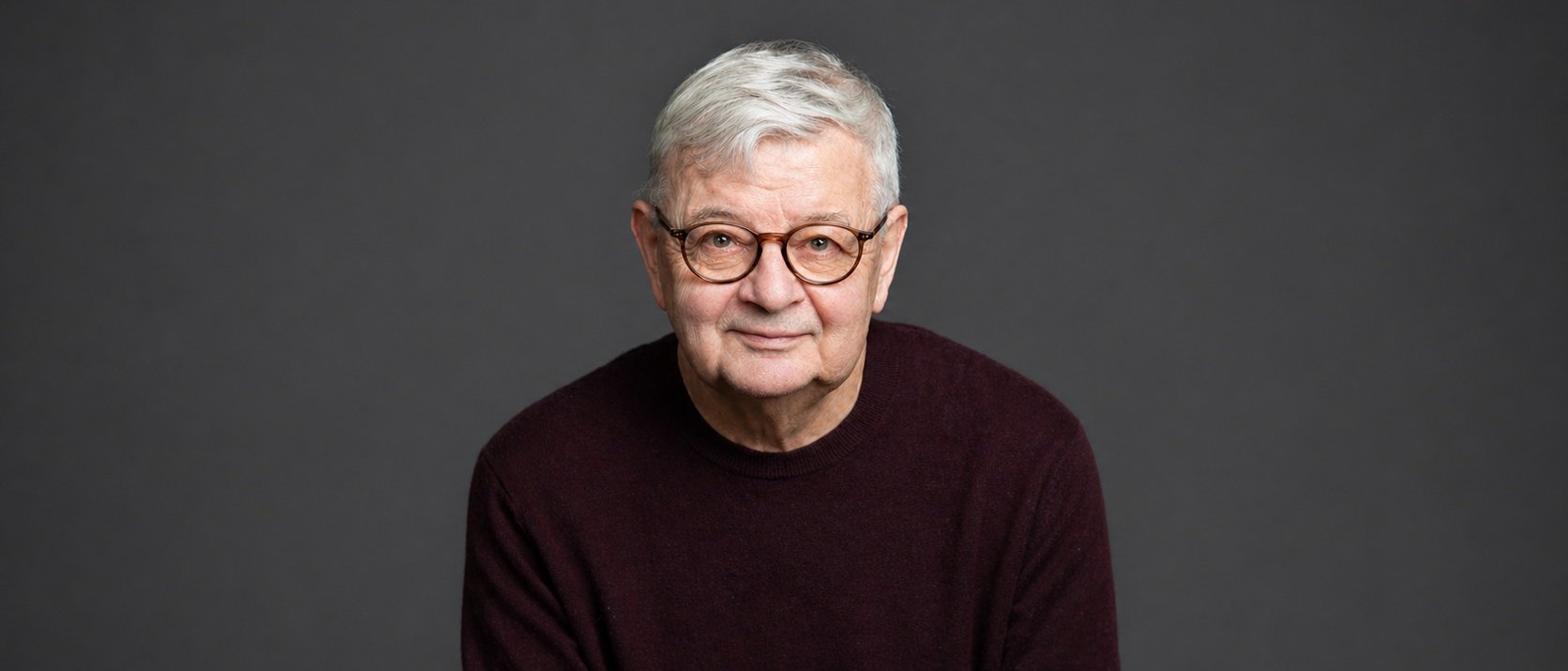 An older man with short gray hair, glasses, and a dark maroon sweater sits in front of a plain dark gray background, looking at the camera and smiling gently.