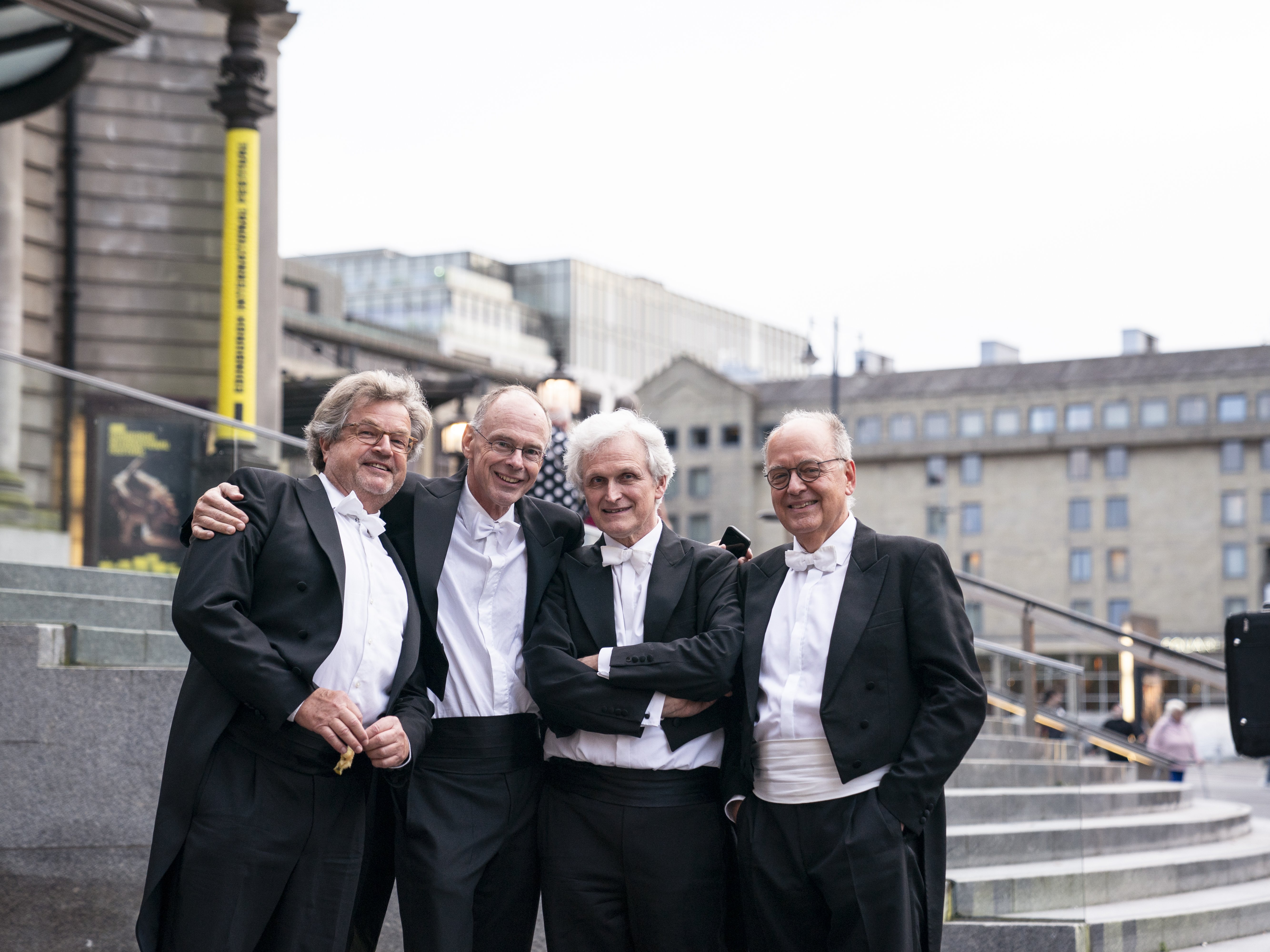 Four older men in formal black and white tuxedos stand close together, smiling, on outdoor steps in an urban setting with buildings in the background.