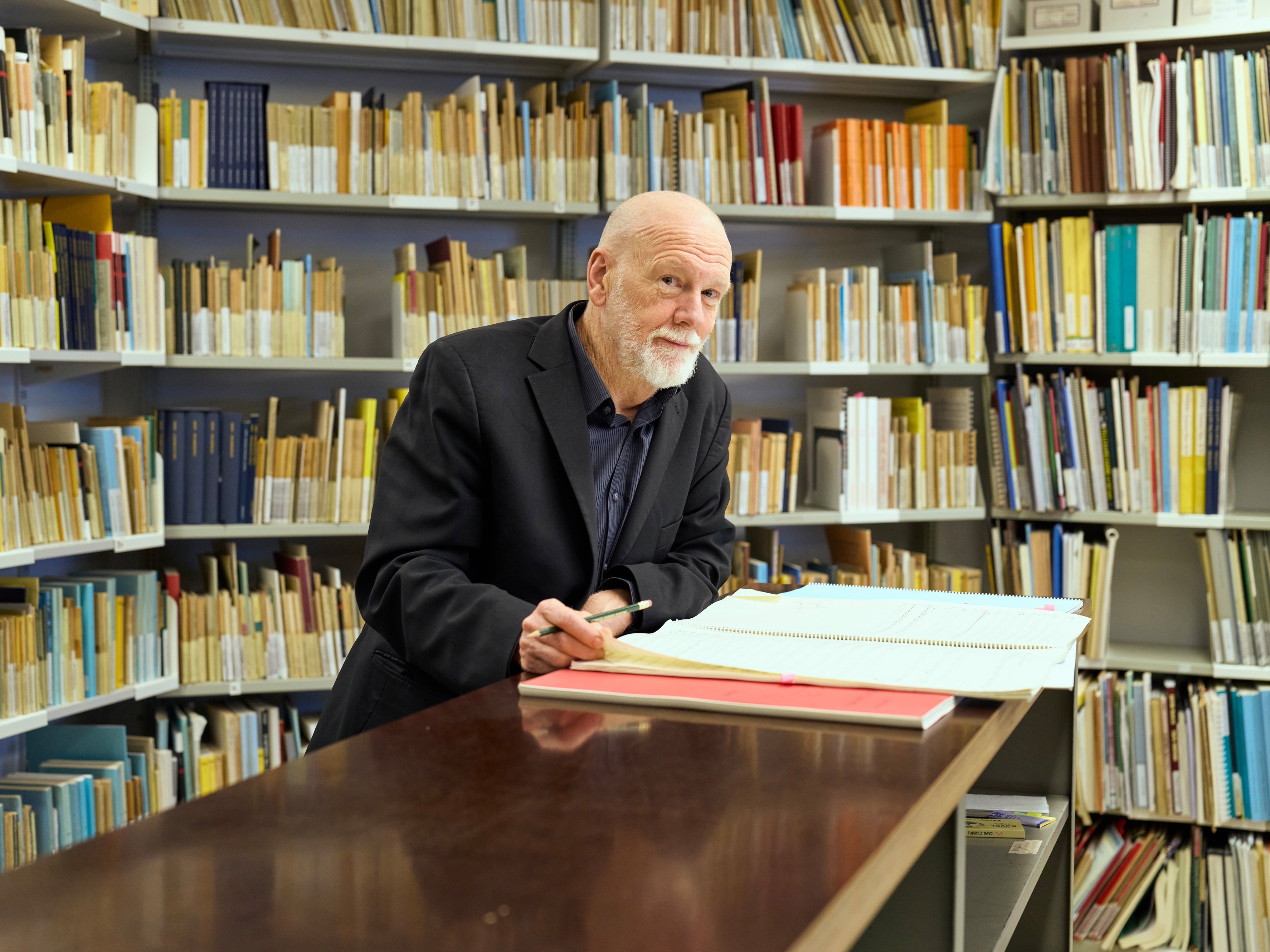 Brett Dean mit Bart steht an einem Schreibtisch in einer Bibliothek und schreibt in ein großes Buch. An den Wänden hinter ihm stehen Regale mit ordentlich geordneten Büchern und Ordnern.