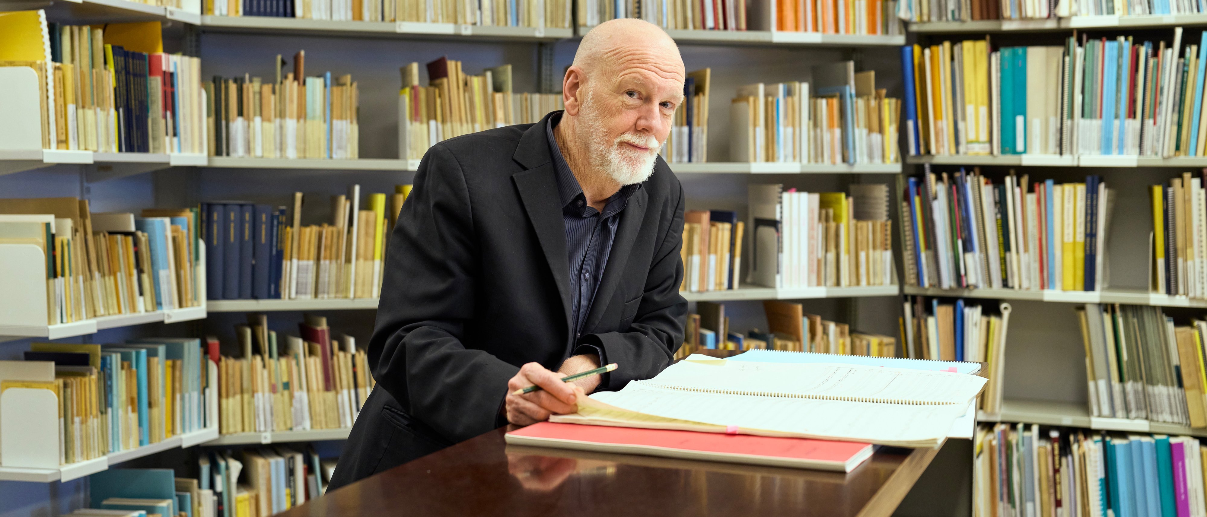 Brett Dean mit Bart steht an einem Schreibtisch in einer Bibliothek und schreibt in ein großes Buch. An den Wänden hinter ihm stehen Regale mit ordentlich geordneten Büchern und Ordnern.