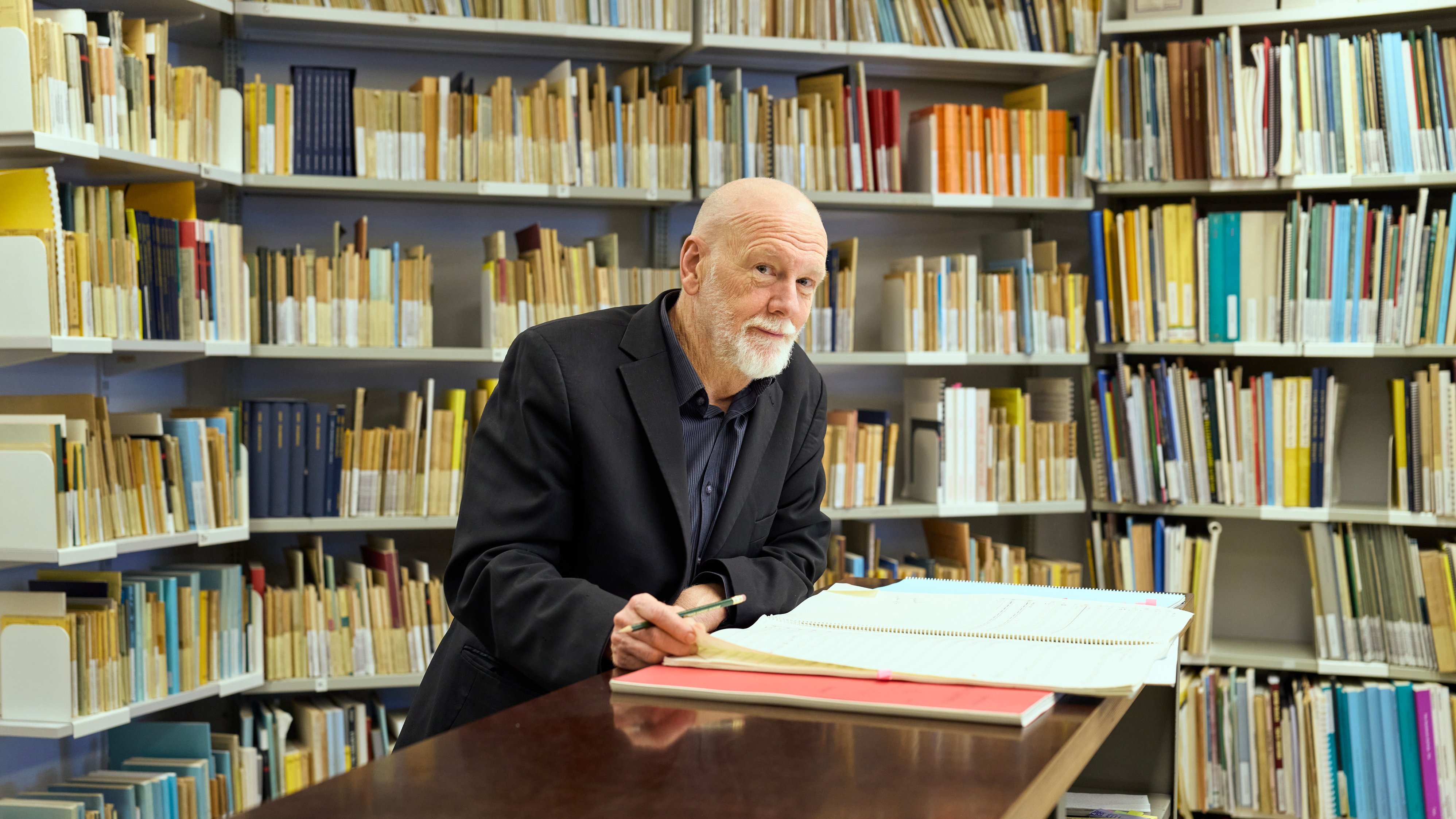 Brett Dean mit Bart steht an einem Schreibtisch in einer Bibliothek und schreibt in ein großes Buch. An den Wänden hinter ihm stehen Regale mit ordentlich geordneten Büchern und Ordnern.
