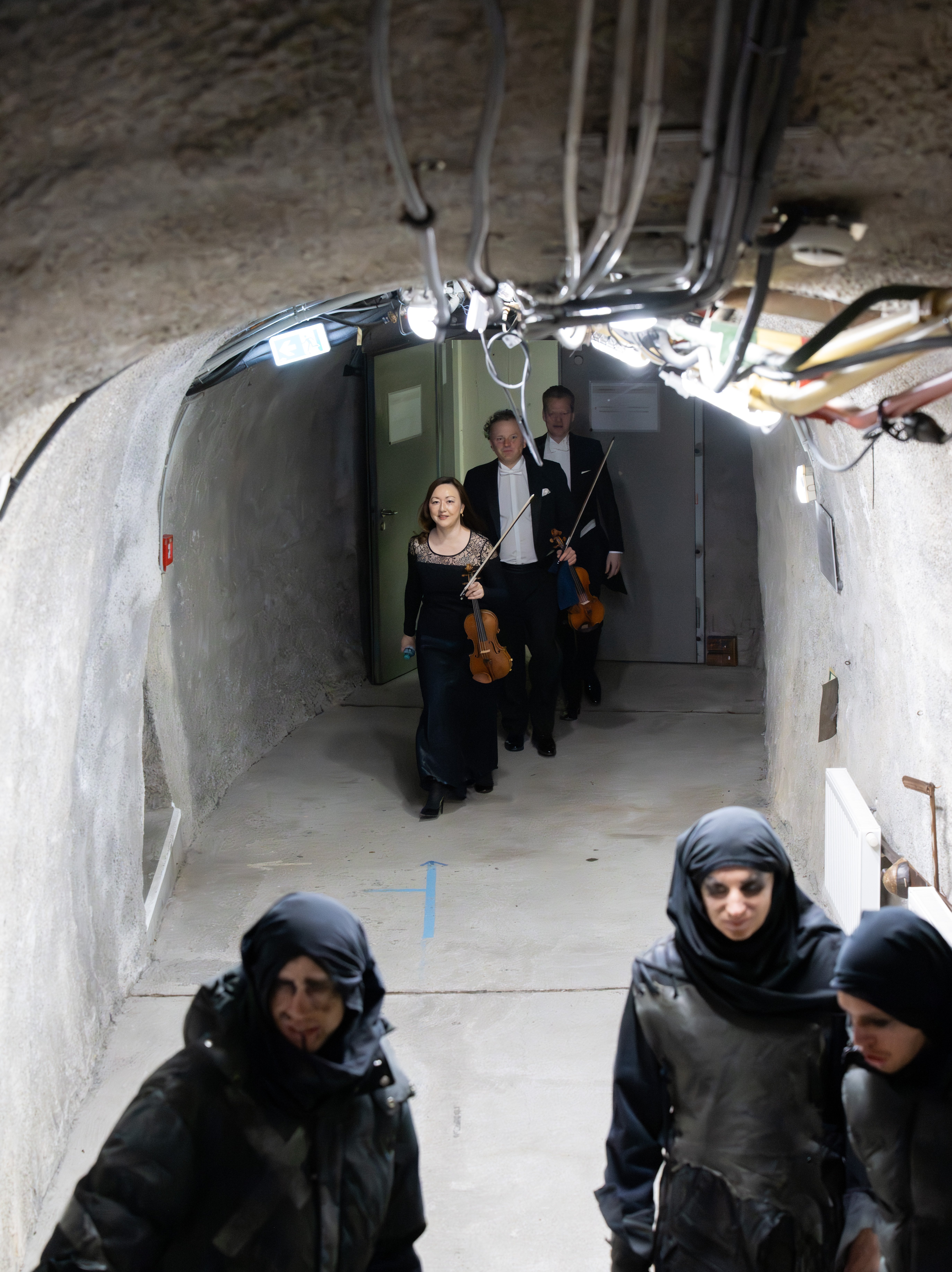 Musiker in förmlicher Kleidung mit Geigen in der Hand gehen durch einen unterirdischen Tunnel, während im Vordergrund Menschen in dunkler taktischer Kleidung mit Kapuzen und Gesichtsbemalung stehen.