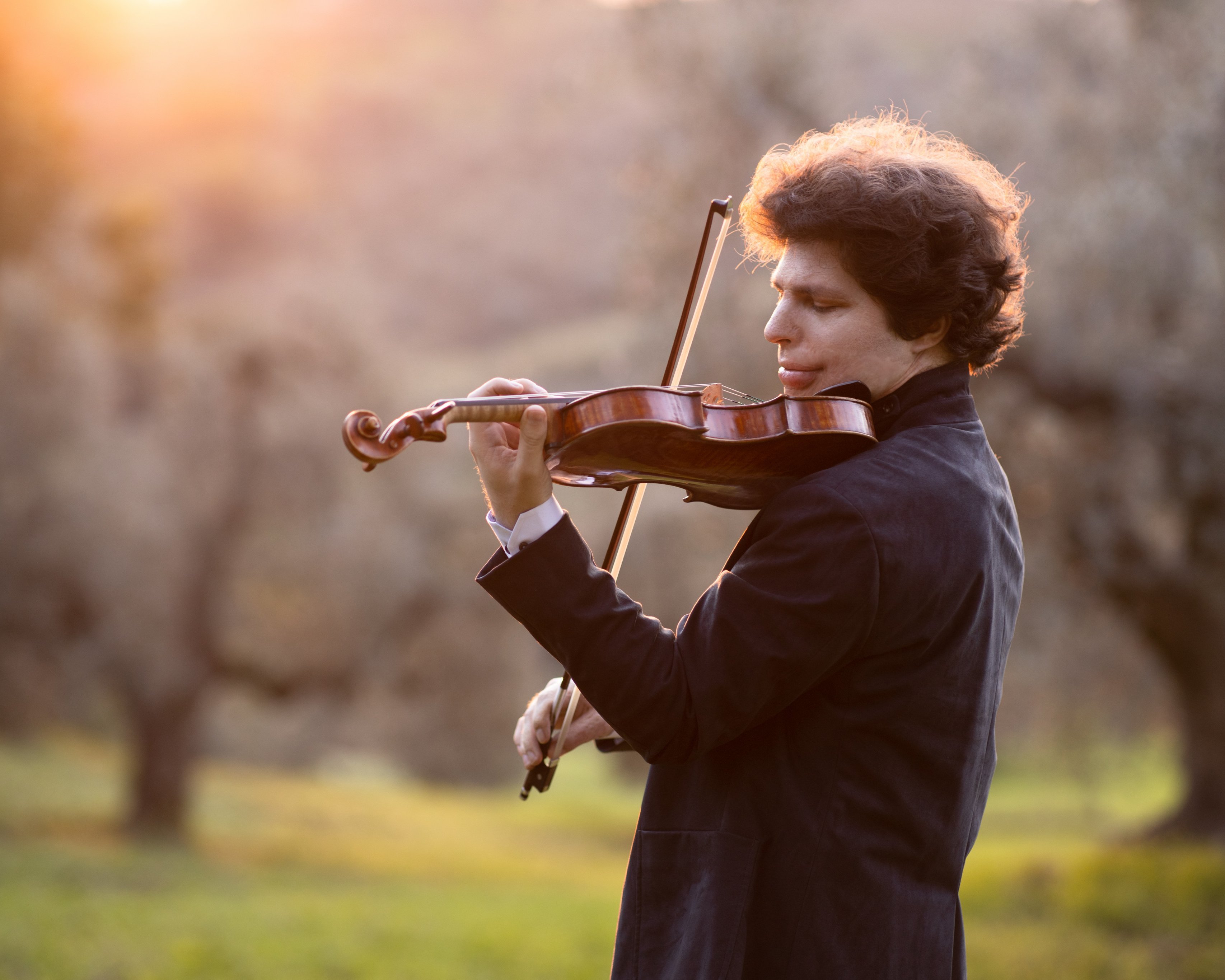 Augustin Hadelich plays the violin outdoors, trees in the background