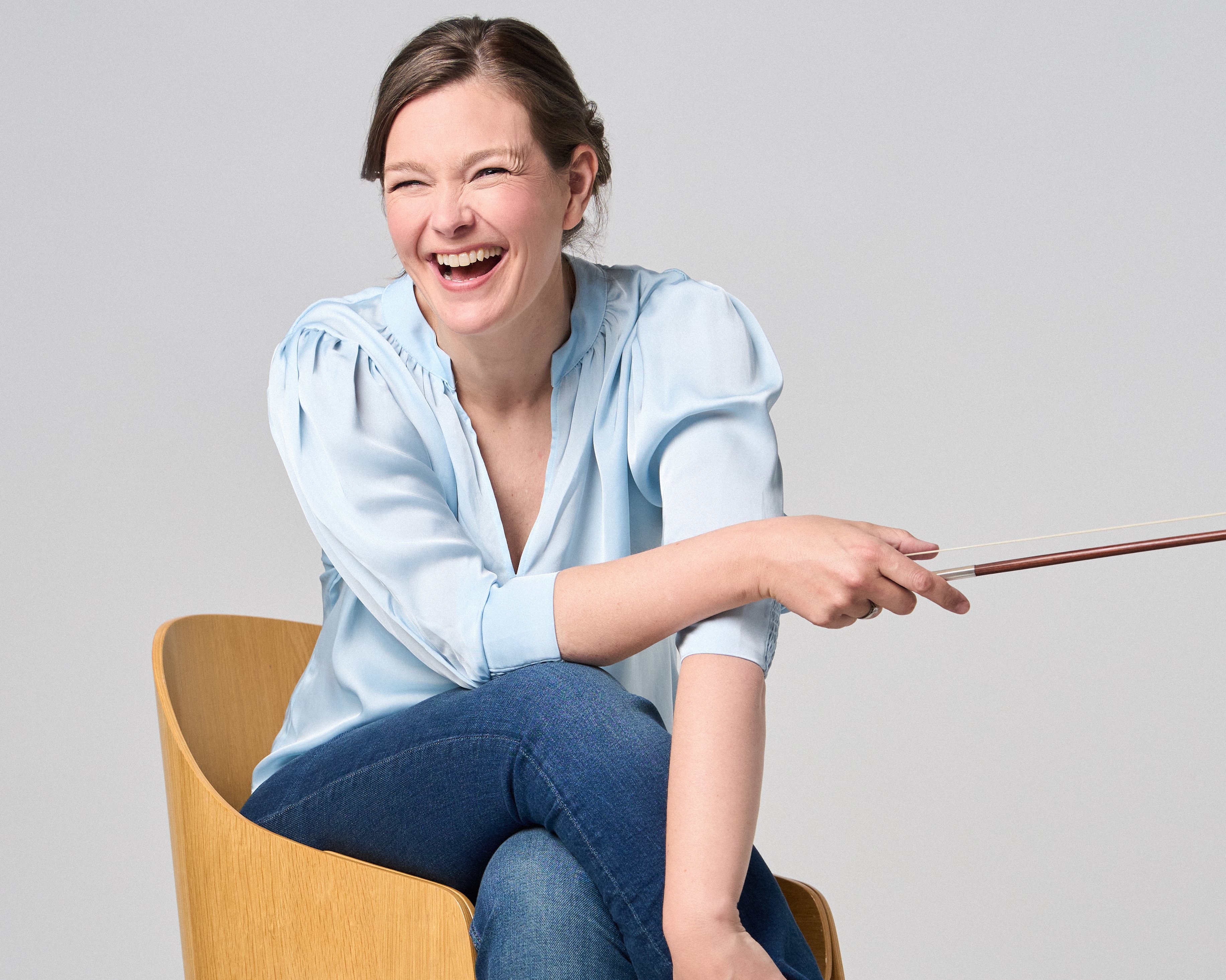 Janine Jansen is sitting on a chair, a violin in her hands. She is wearing jeans and a light blue blouse and looks into the camera with a smile.