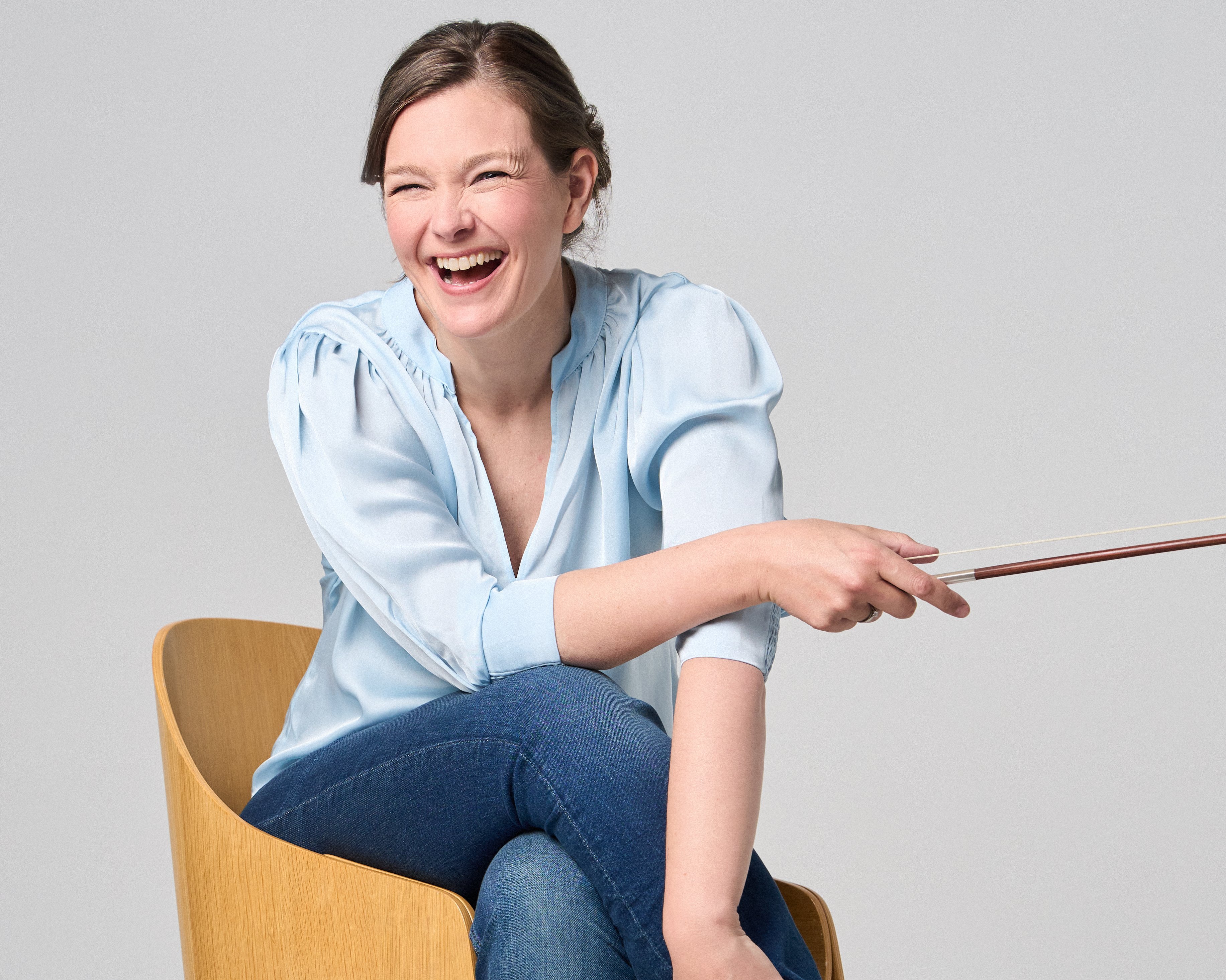 Janine Jansen sitzt auf einem Stuhl, eine Violine in den Händen. Sie trägt Jeans und eine hellblaue Bluse und blickt lächelnd in die Kamera.