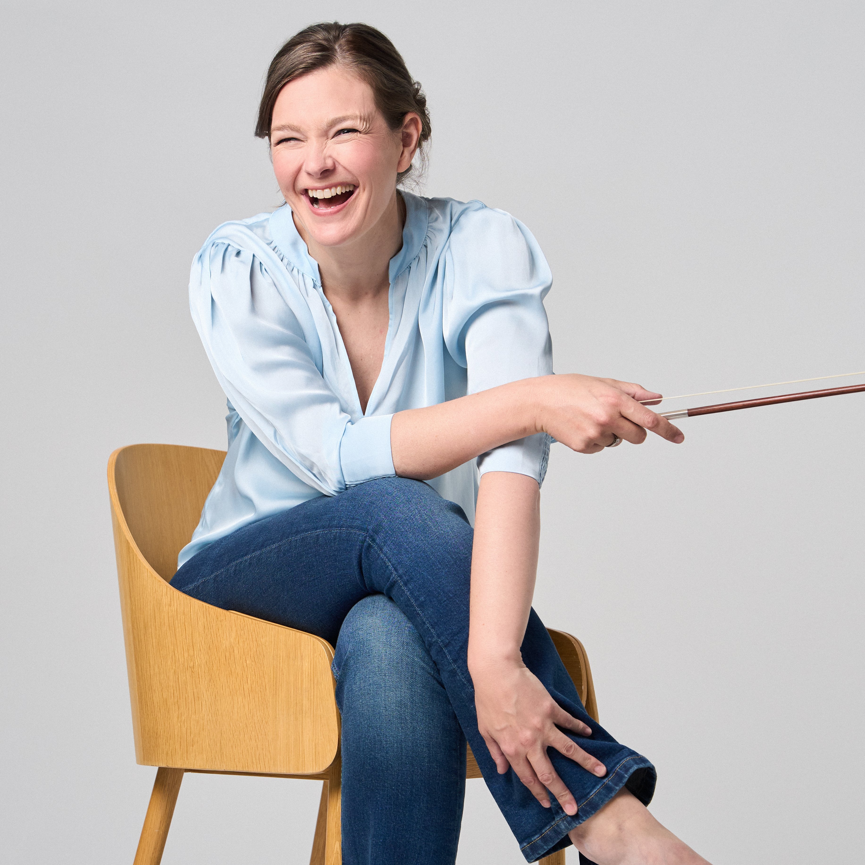 Janine Jansen is sitting on a chair, a violin in her hands. She is wearing jeans and a light blue blouse and looks into the camera with a smile.