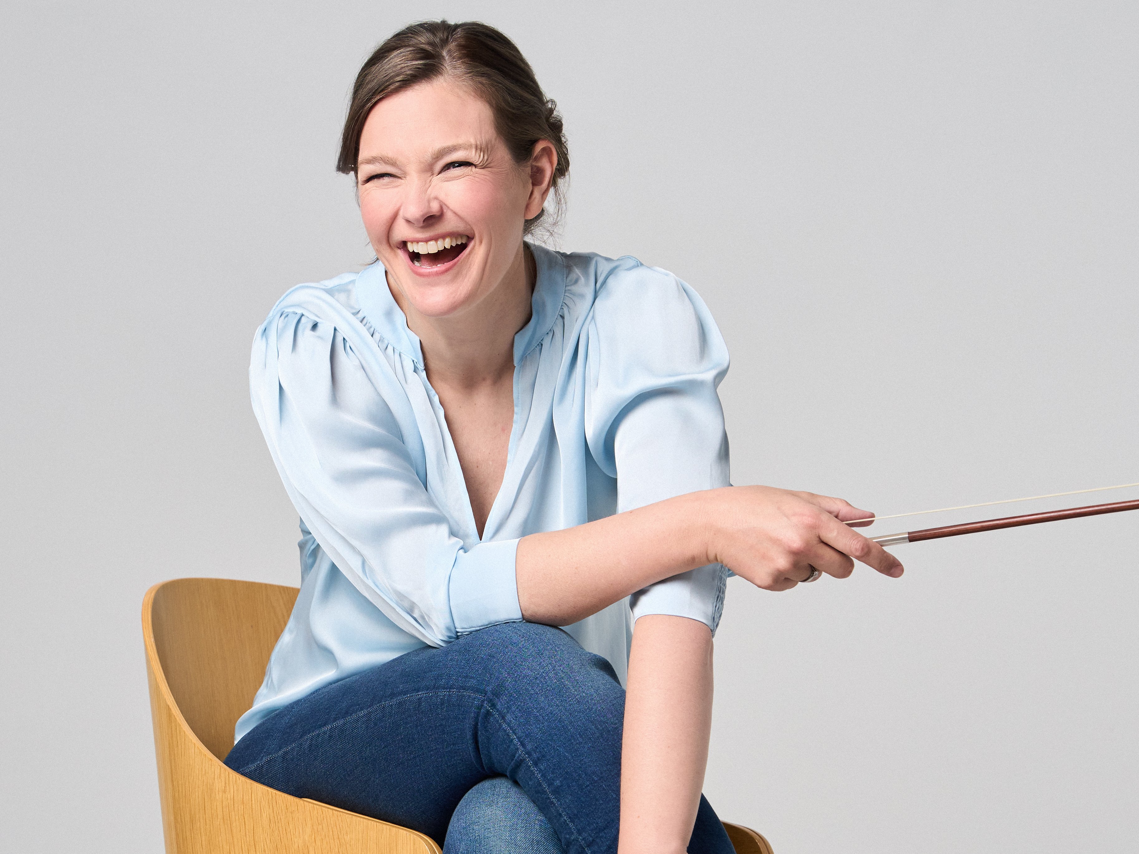 Janine Jansen sitzt auf einem Stuhl, eine Violine in den Händen. Sie trägt Jeans und eine hellblaue Bluse und blickt lächelnd in die Kamera.