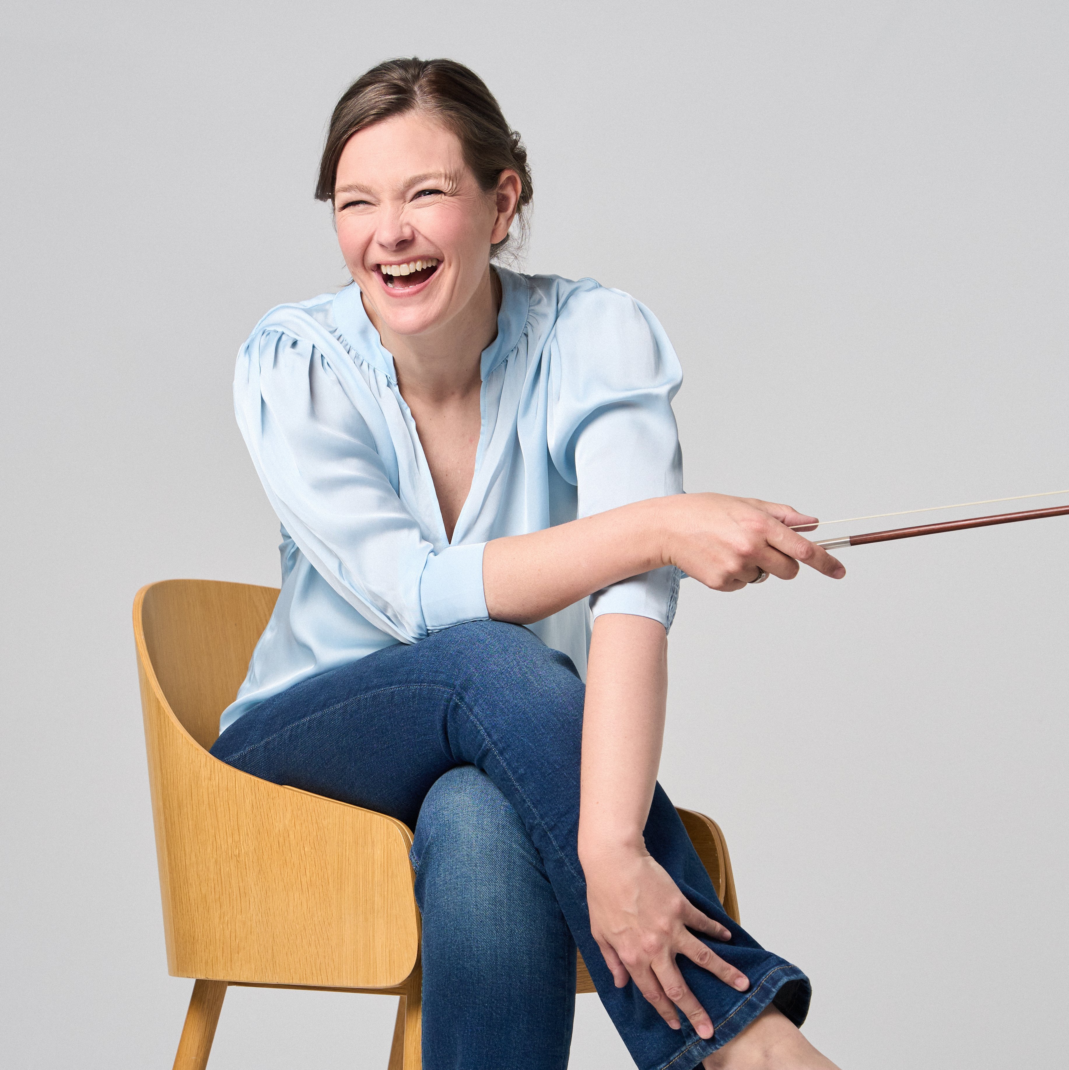 Janine Jansen sitzt auf einem Stuhl, eine Violine in den Händen. Sie trägt Jeans und eine hellblaue Bluse und blickt lächelnd in die Kamera.