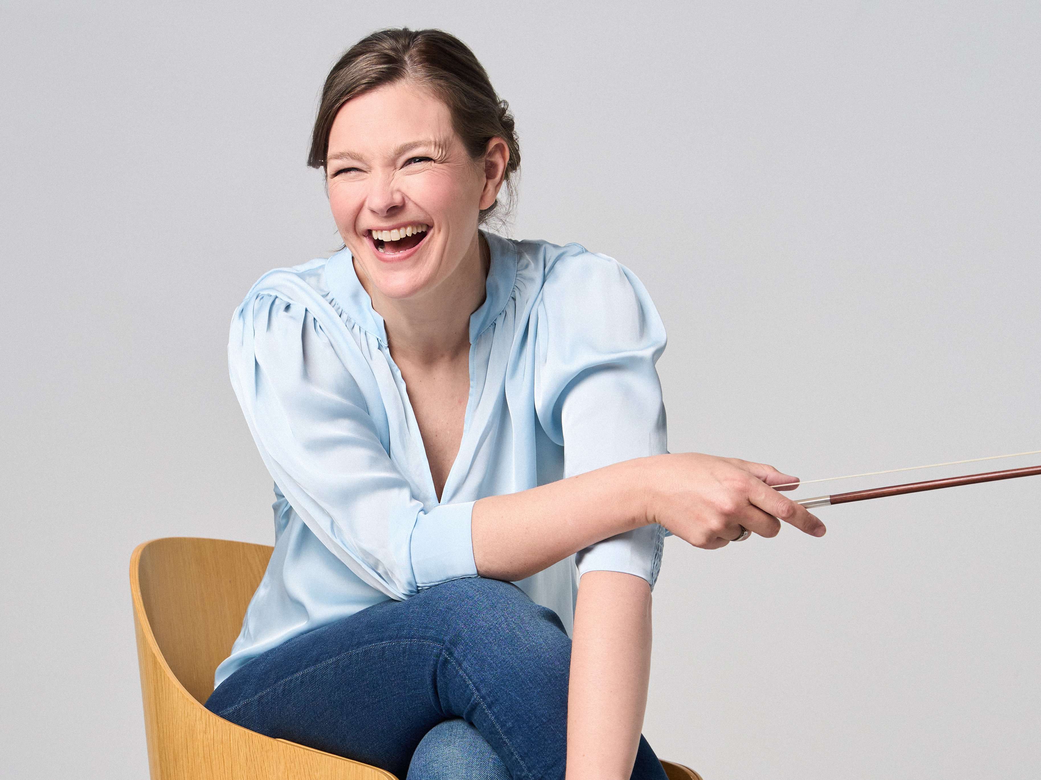 Janine Jansen is sitting on a chair, a violin in her hands. She is wearing jeans and a light blue blouse and looks into the camera with a smile.