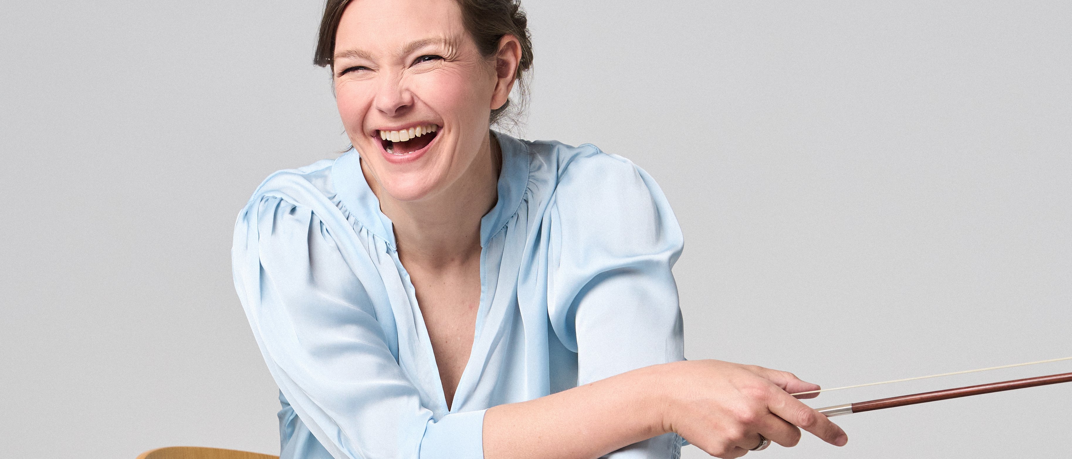 Janine Jansen is sitting on a chair, a violin in her hands. She is wearing jeans and a light blue blouse and looks into the camera with a smile.