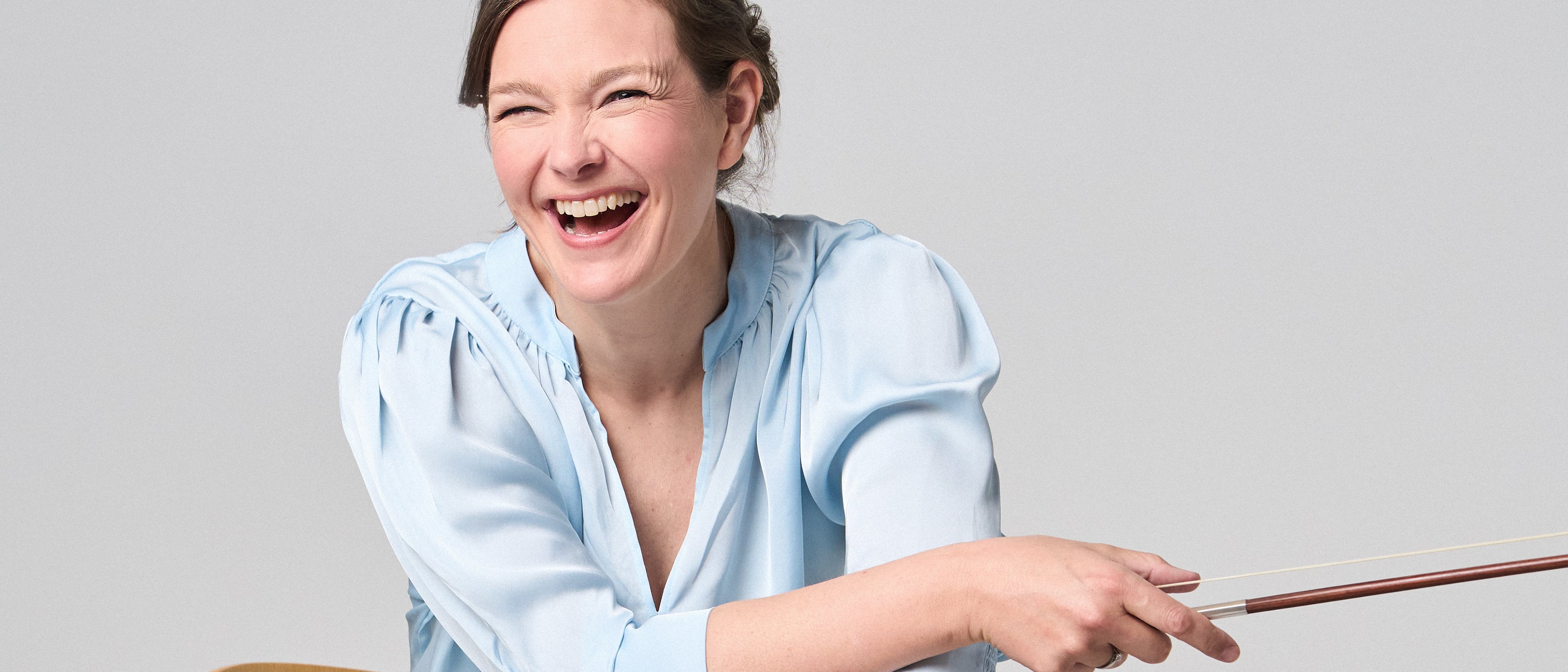 Janine Jansen is sitting on a chair, a violin in her hands. She is wearing jeans and a light blue blouse and looks into the camera with a smile.