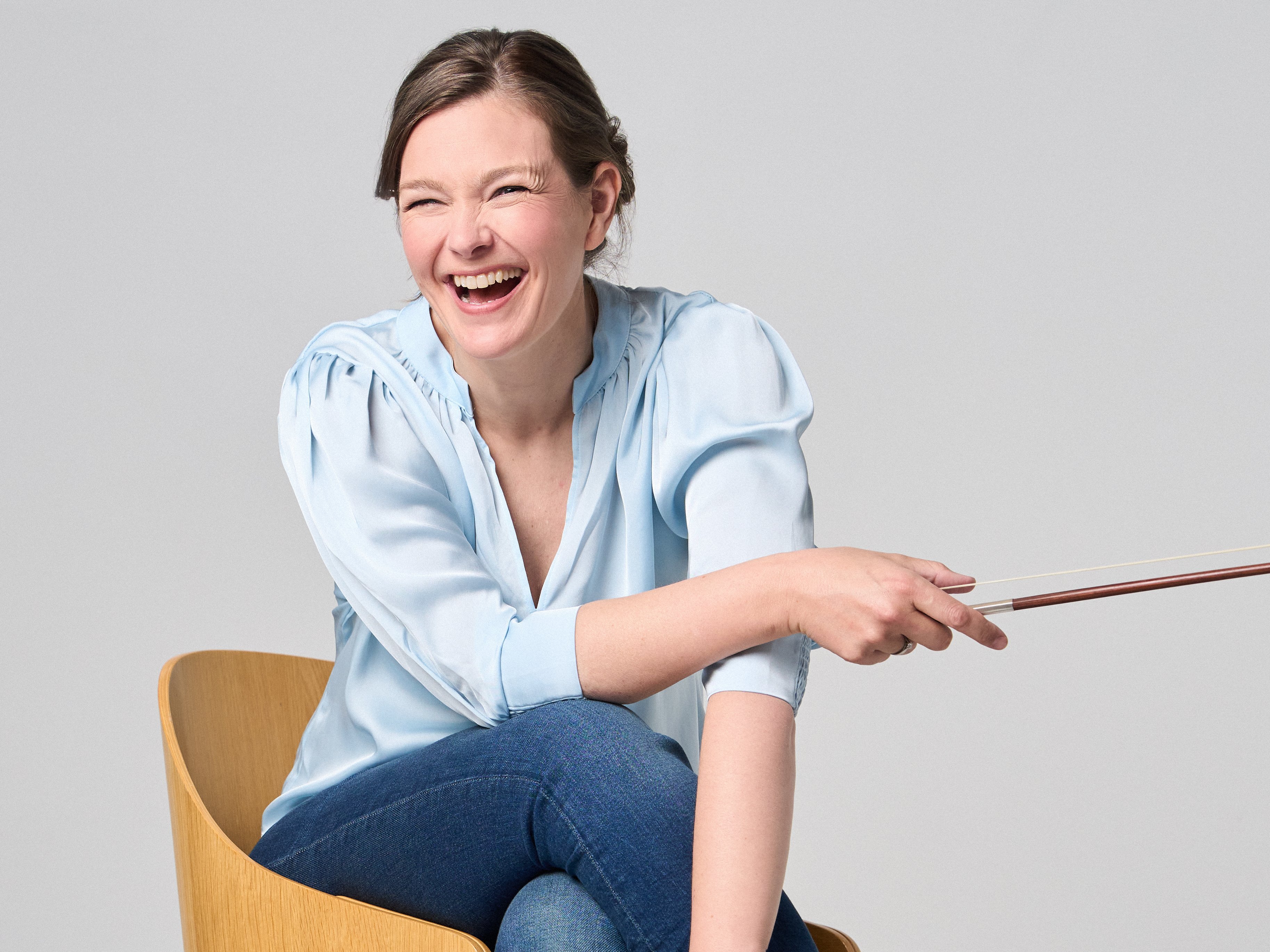 Janine Jansen is sitting on a chair, a violin in her hands. She is wearing jeans and a light blue blouse and looks into the camera with a smile.
