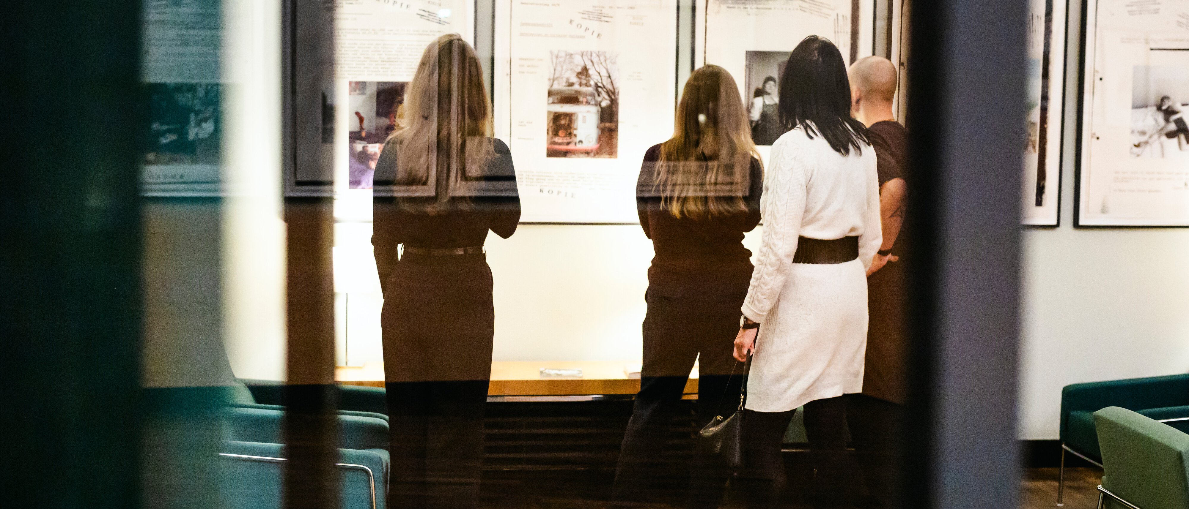 Vier Personen stehen in einer Kunstgalerie oder einem Museum und betrachten gerahmte Bilder oder Dokumente, die an einer Wand ausgestellt sind. Die Szene wird durch eine Glaswand betrachtet, die eine subtile Reflexion erzeugt.