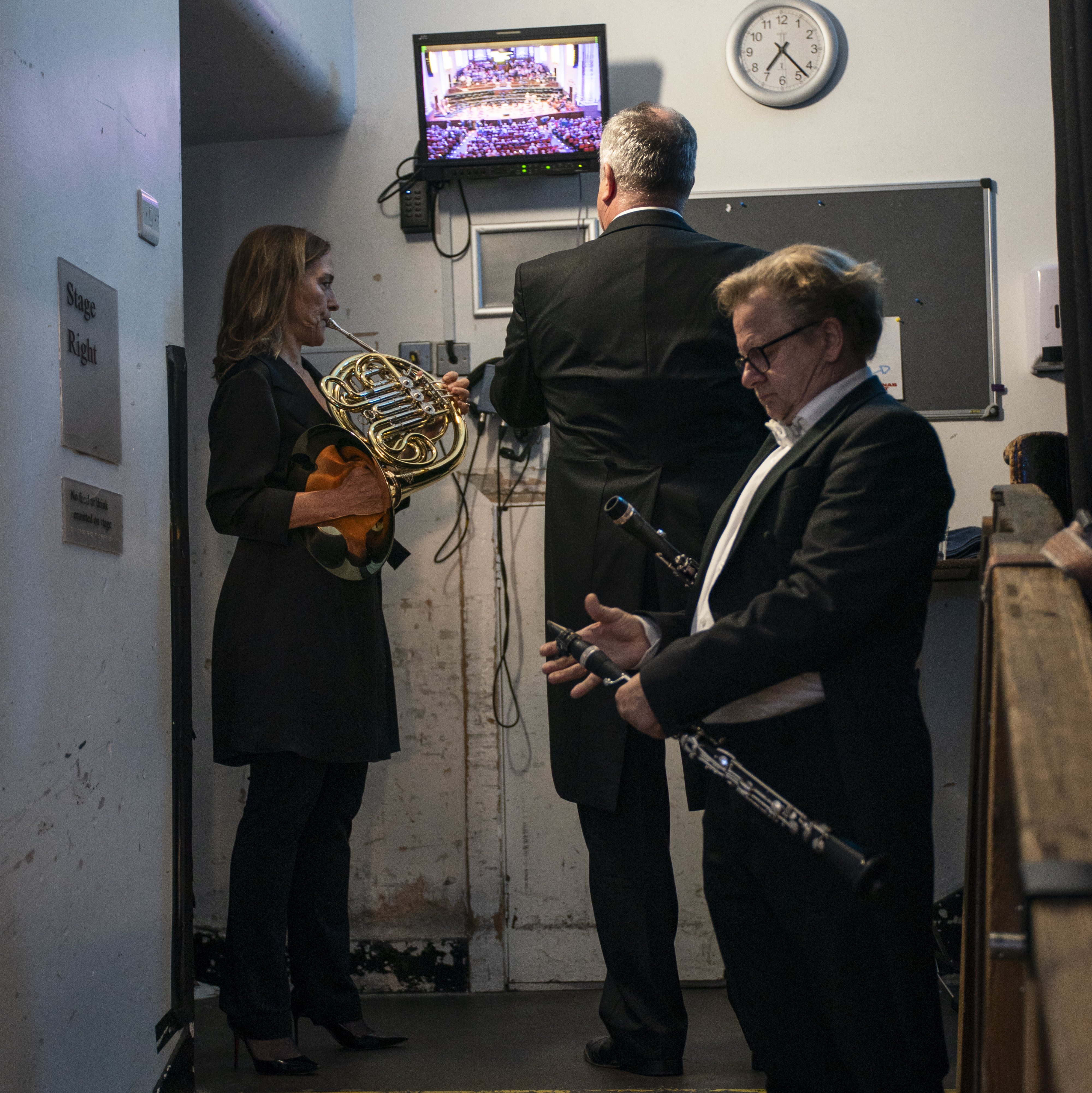 Three musicians in formal wear wait backstage. One holds a French horn, another checks a monitor showing an orchestra, and the third holds a clarinet. A clock and backstage signs are on the wall.