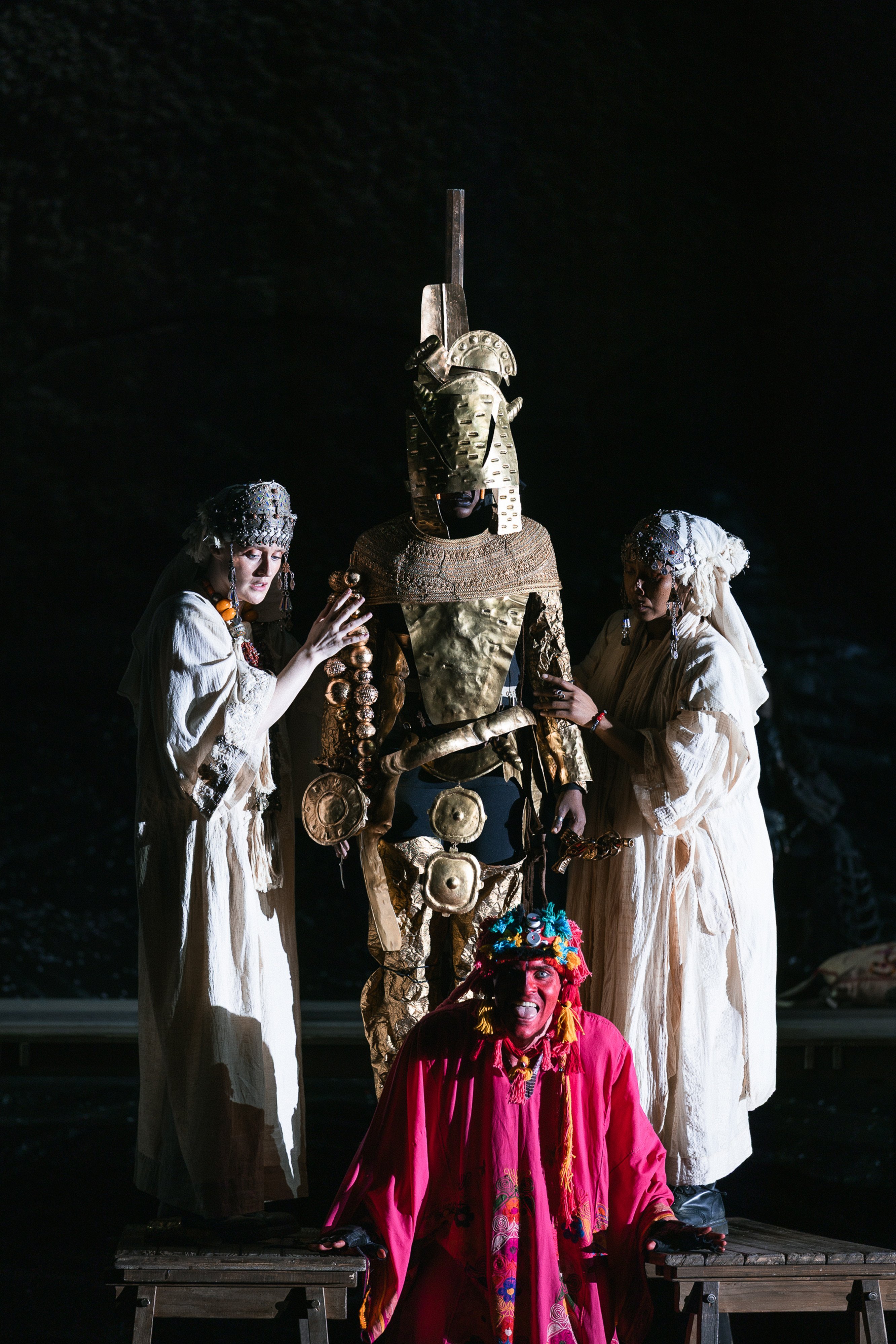 Three people in white robes stand beside a person in elaborate golden armor and headdress, while a fourth person in a bright red costume with a colorful headdress kneels in front. The scene is dimly lit.