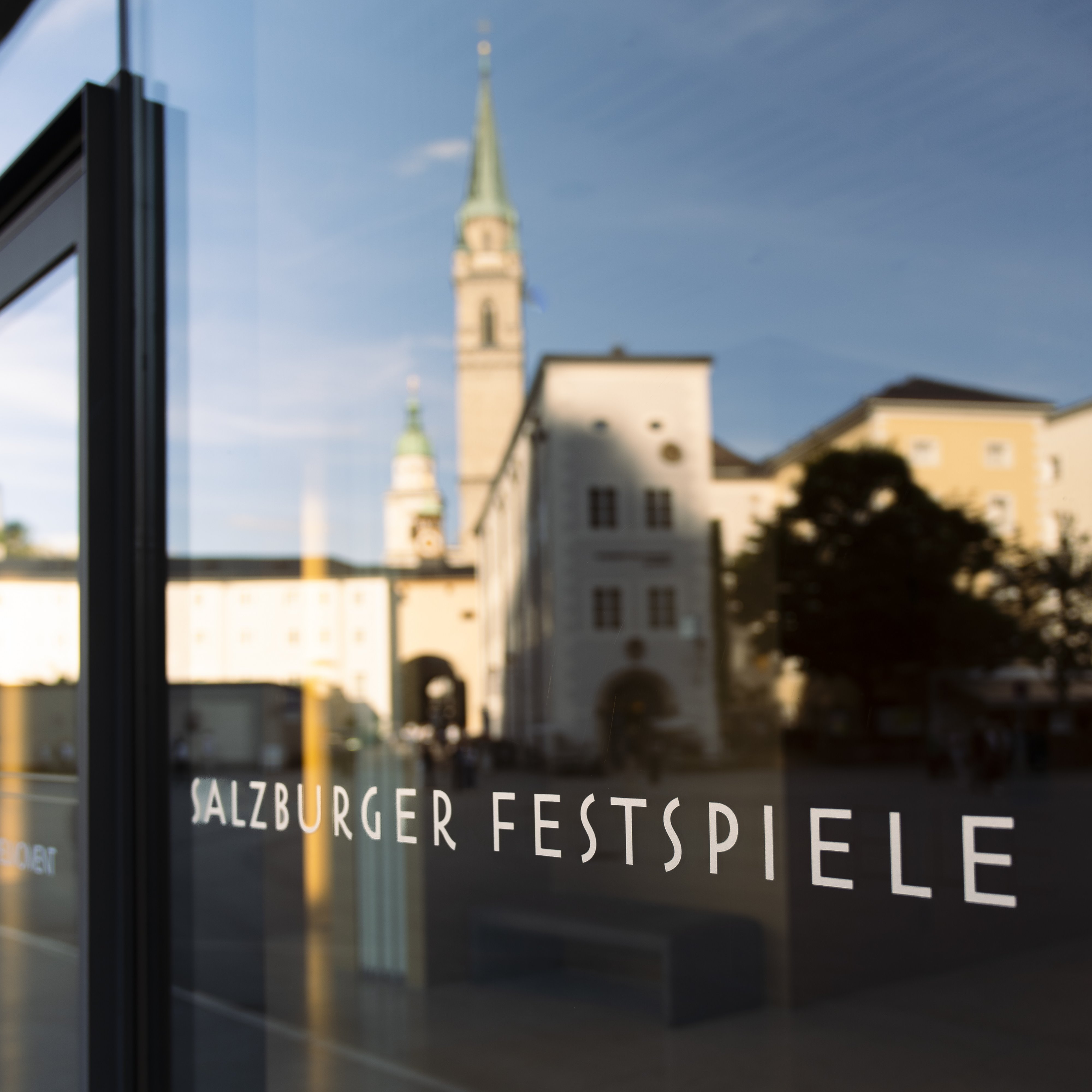 Die Spiegelung eines Kirchturms und von Gebäuden in einer Glastür, auf der der Schriftzug „Salzburger Festspiele» zu sehen ist.
