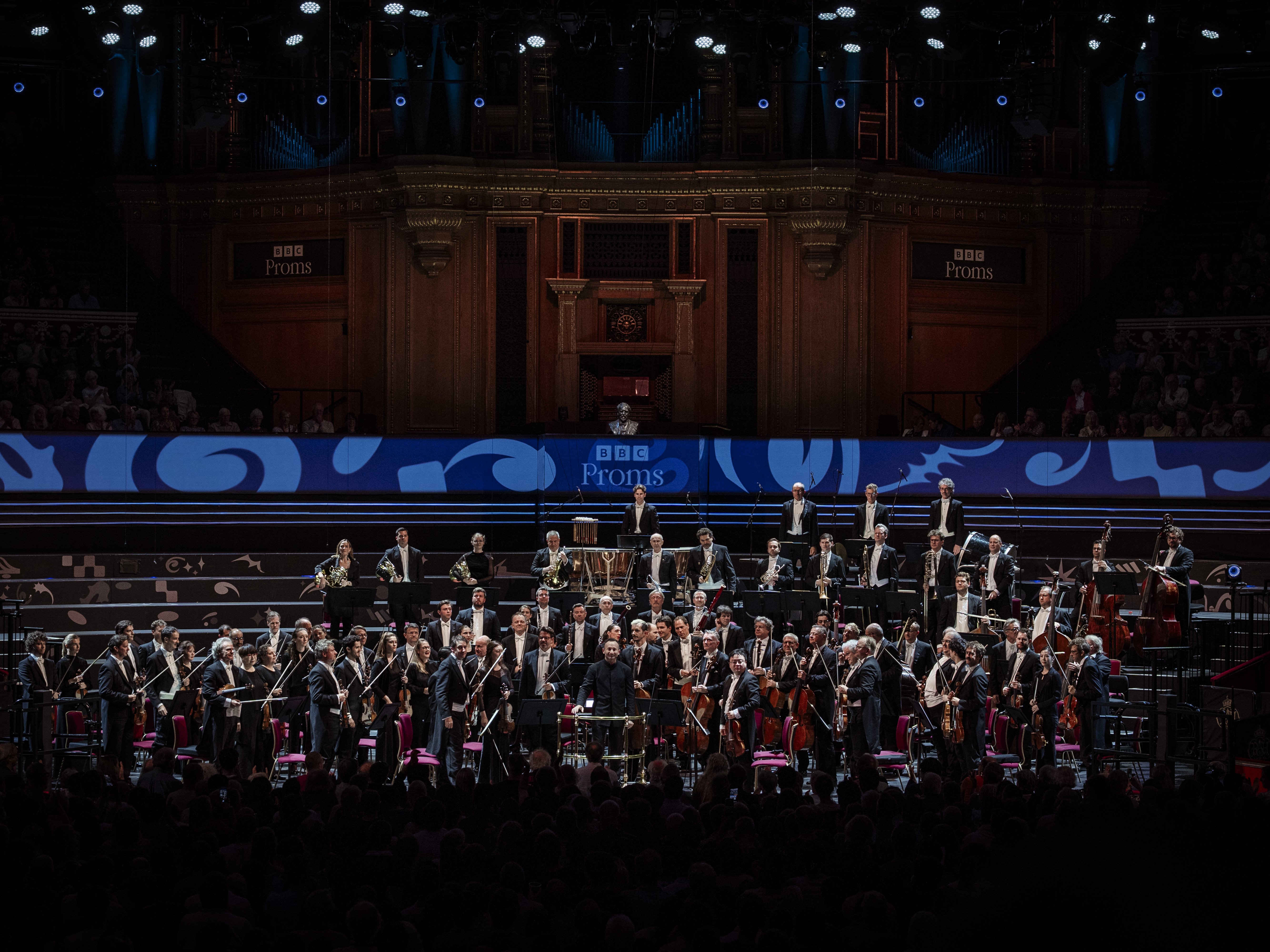 Auf der Bühne der BBC Proms spielt ein vollbesetztes Orchester, wobei die Musiker in einem prächtigen Saal unter Konzertbeleuchtung verschiedene Instrumente spielen.