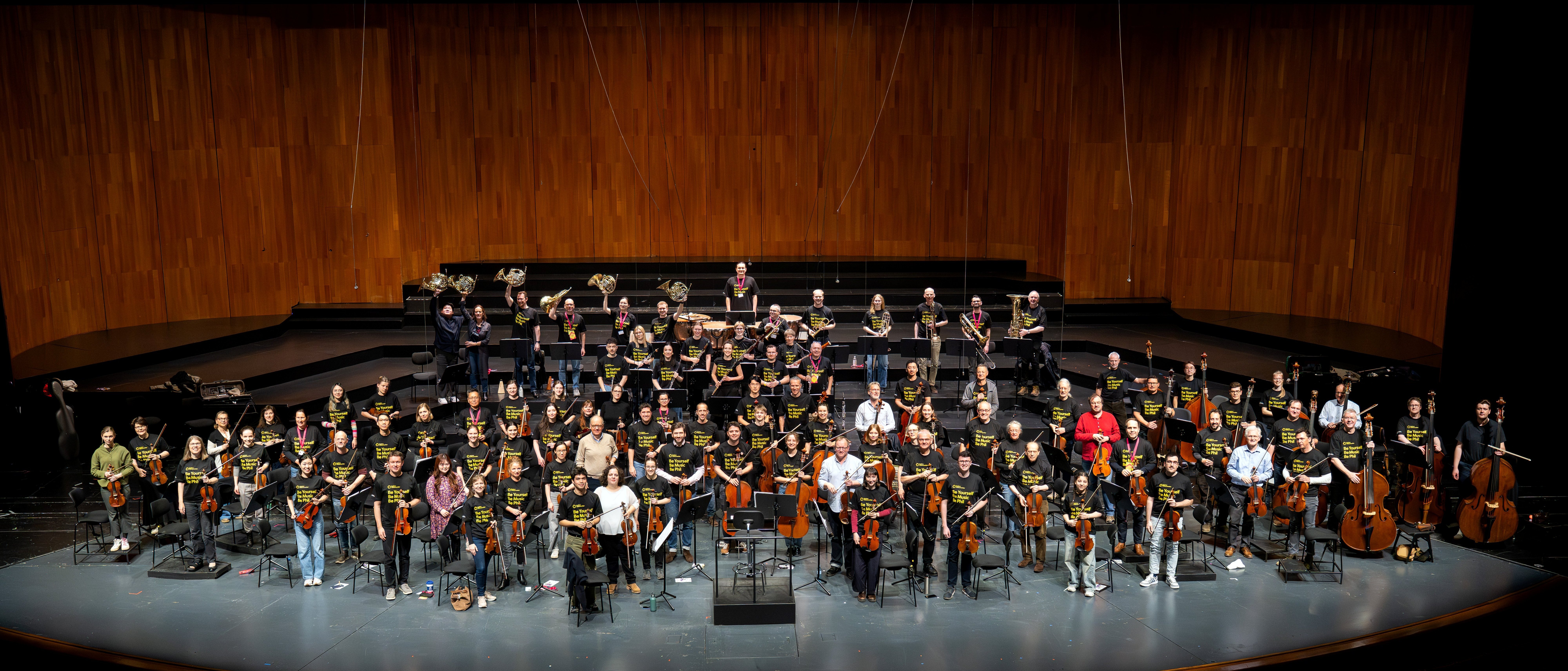 A large orchestra on a stage with string, brass, and woodwind players in matching shirts against a wooden backdrop.